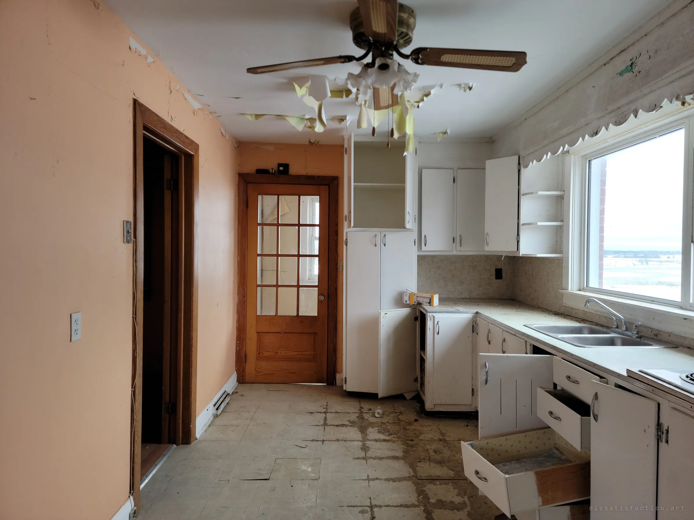 The kitchen seen lengthwise, peach walls and a glazed wooden door at the end, while paper peels from the ceiling around a ceiling fan. Open drawers and a stained tile floor show the slow decay.