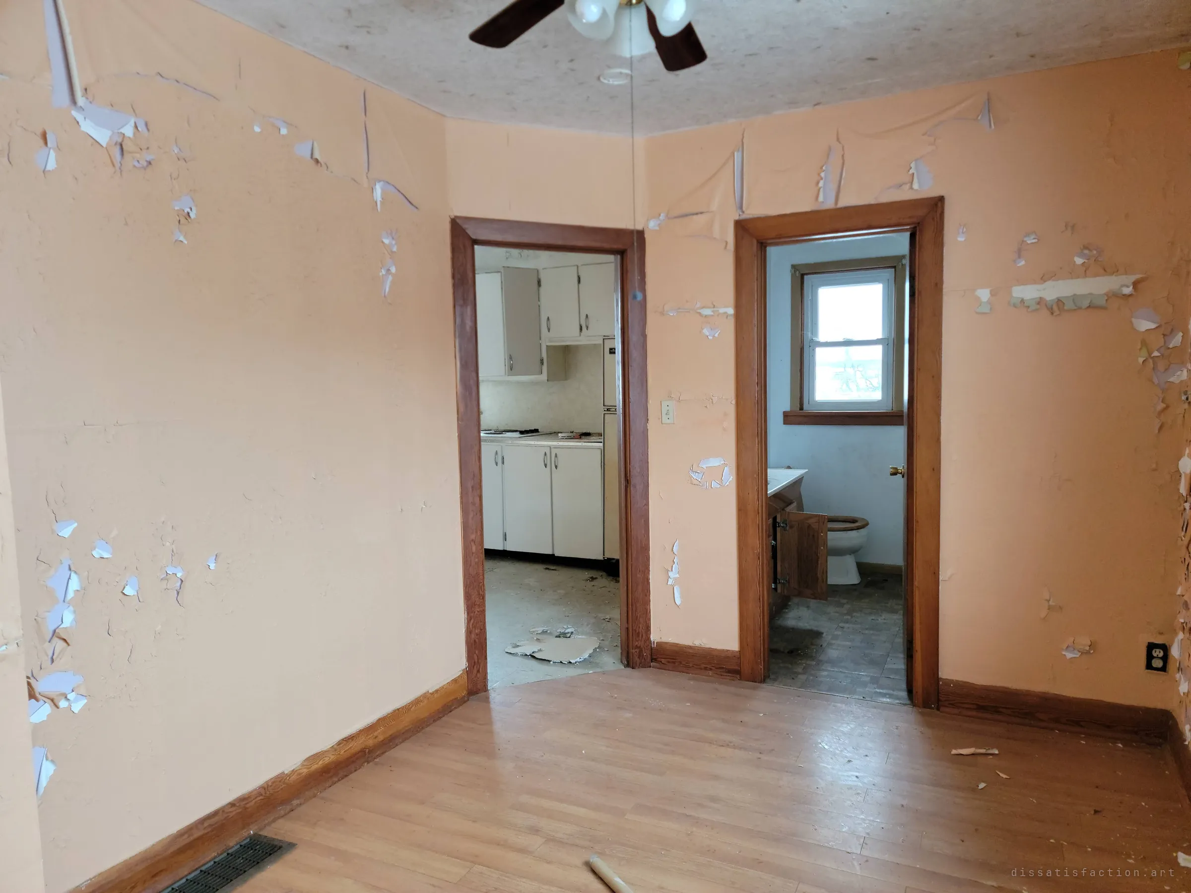 Two wood-trimmed doorways in a peach room with flaking paint lead into the kitchen and a small bathroom. The layout of the house unfolds from this one spot.