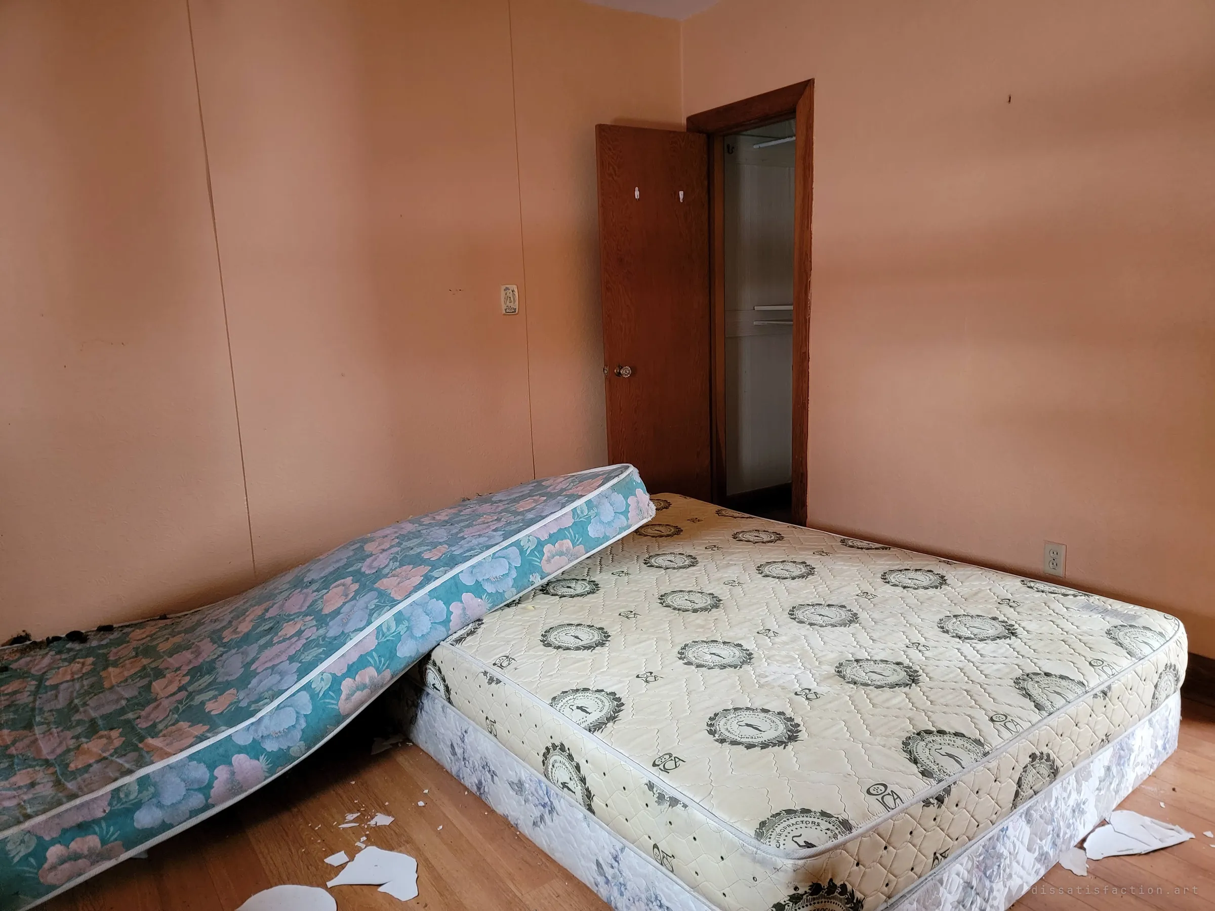 Two mattresses lie tumbled on the floor of a peach bedroom, one slumped over the other, beside an open closet door. Flakes of ceiling litter the wooden floor.