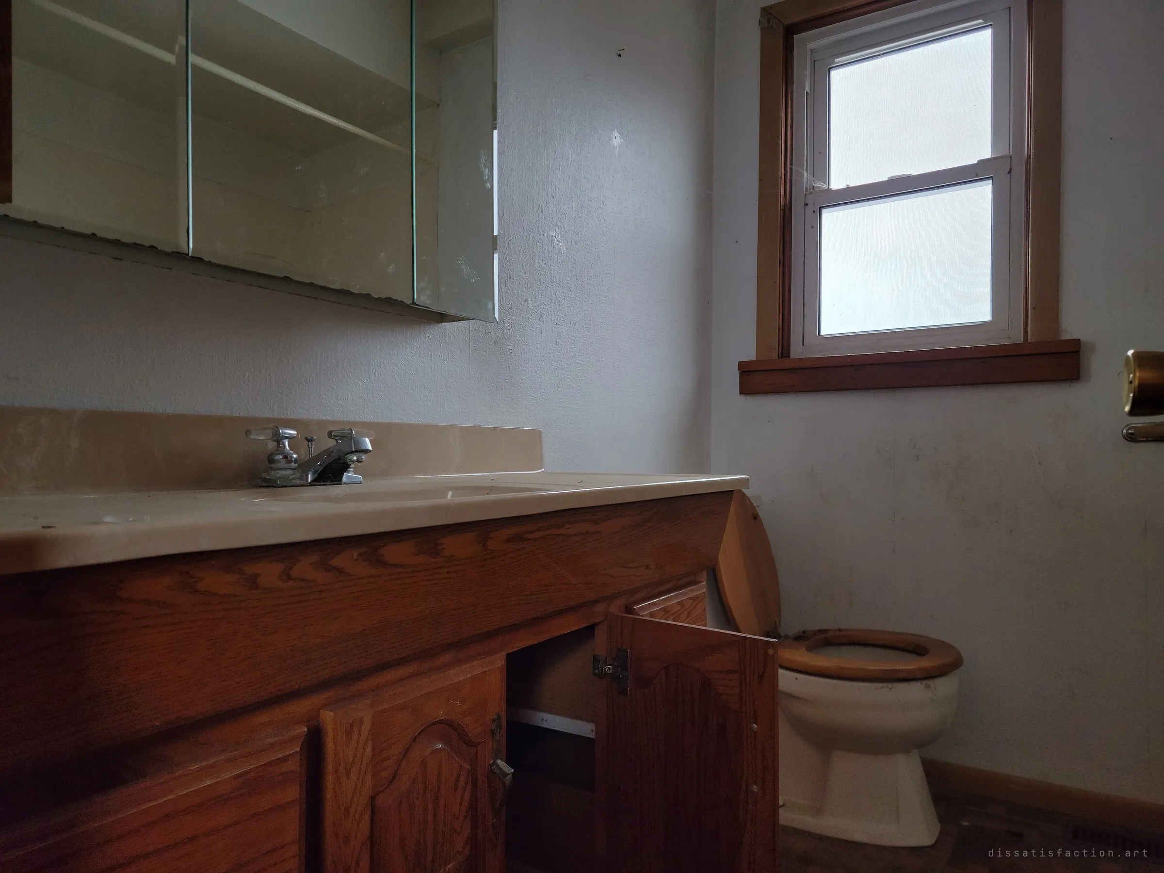 A low view across an oak bathroom vanity toward a toilet with a wooden seat under a frosted window. The mirrored cabinet above hangs open and empty.