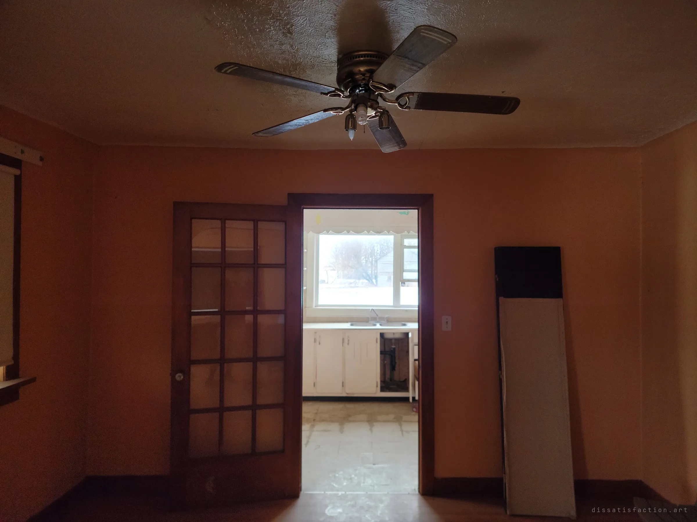 From a dim room with a brass ceiling fan, a doorway beside a glazed French door opens onto the bright kitchen and its window. The contrast pulls the eye through the house.