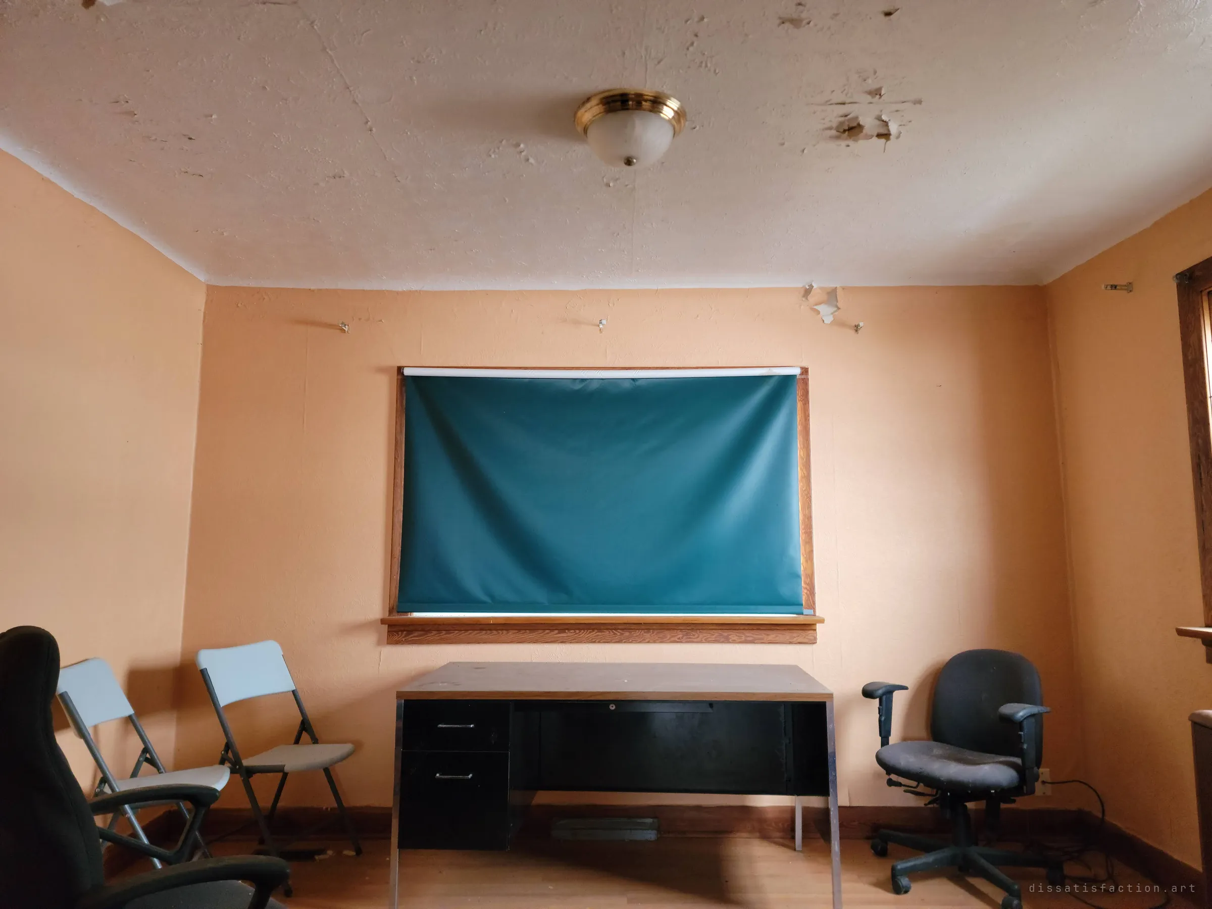 The office the album is named for: a steel desk under a drawn teal blind, flanked by folding chairs and a worn swivel chair. The furniture was left in place as if a meeting might resume.