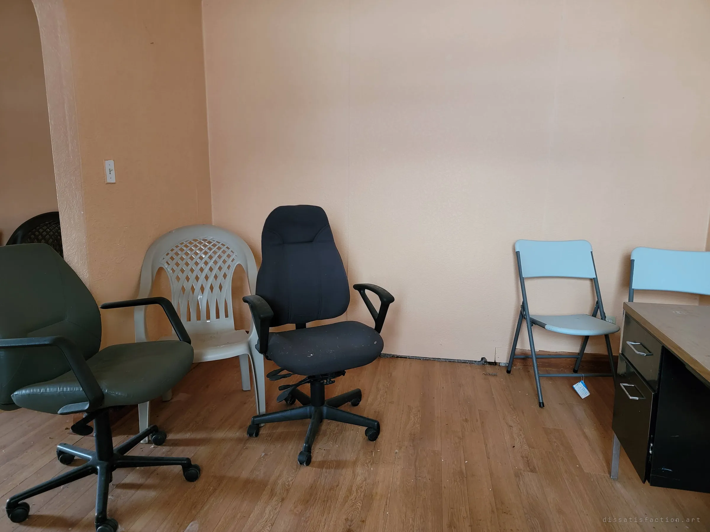 A row of mismatched chairs, office swivel seats, a plastic patio chair and folding chairs, lined up against a peach wall beside a steel desk. The furniture is left as if waiting for a meeting that never happened.