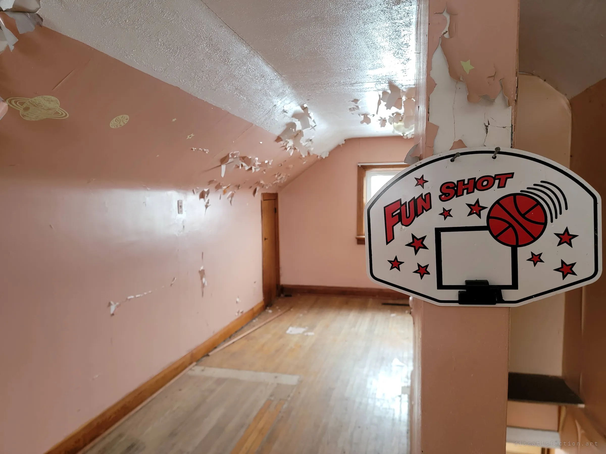 Close on a toy basketball backboard hung on a pink post, the long attic room behind it strewn with paint flakes and glow-in-the-dark planet stickers on the sloped wall. Small traces of whoever played up here.