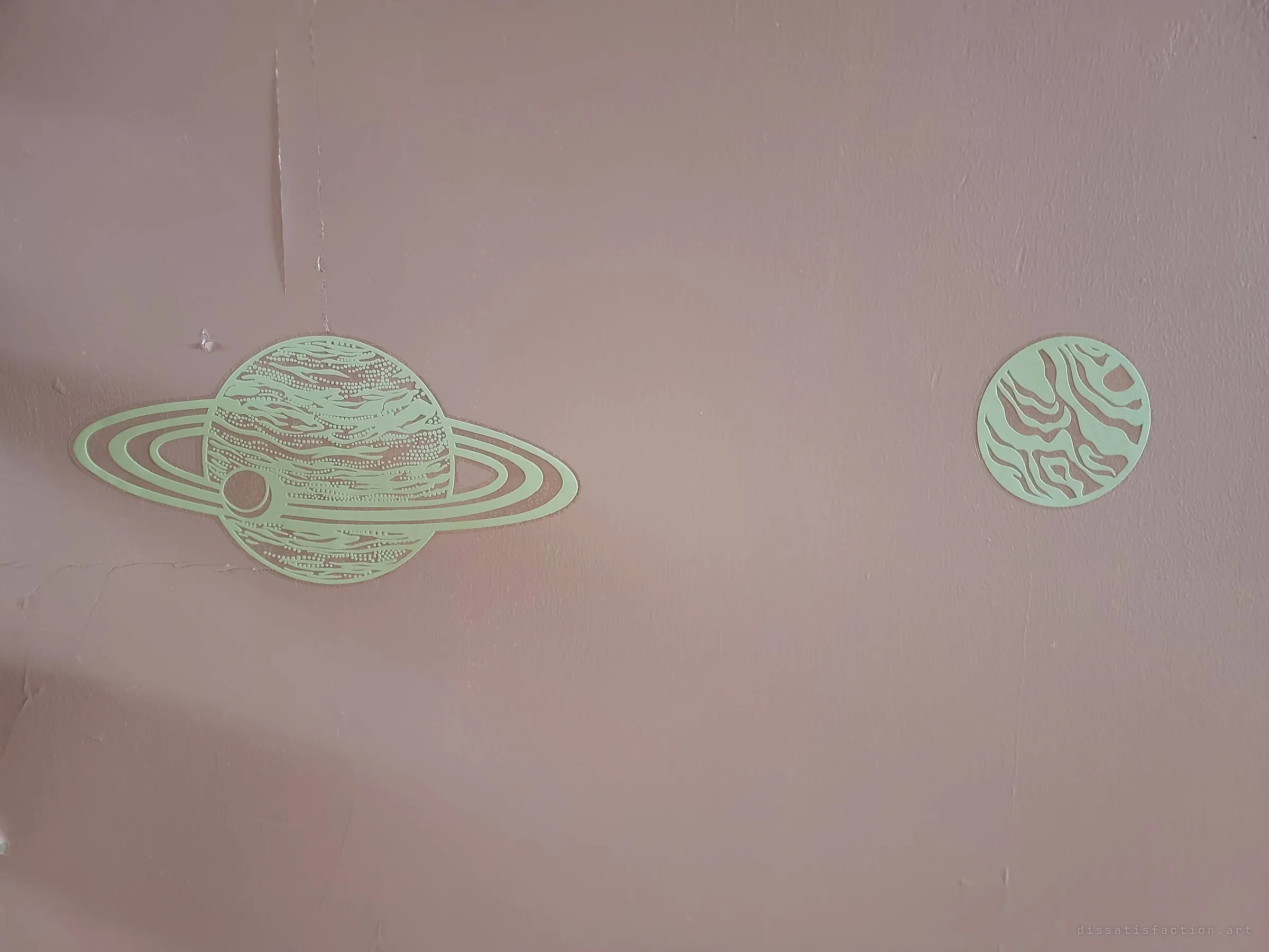 Two glow-in-the-dark planet decals, a ringed planet and a banded moon, stuck to a cracked pink wall. A child's night sky left behind in the dark.