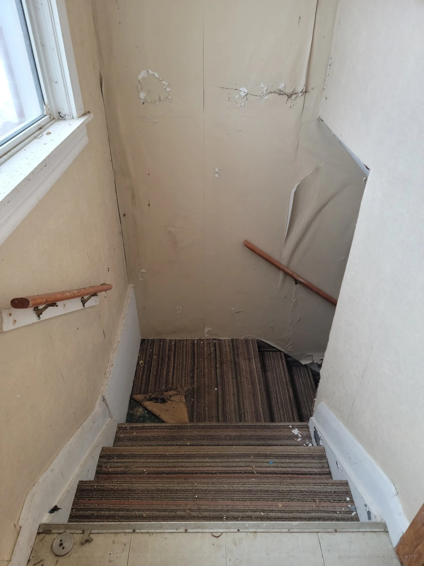 Looking down a narrow carpeted staircase between cream walls, a wooden handrail on each side and wallpaper peeling off the landing. The window light fades as the steps turn out of sight.