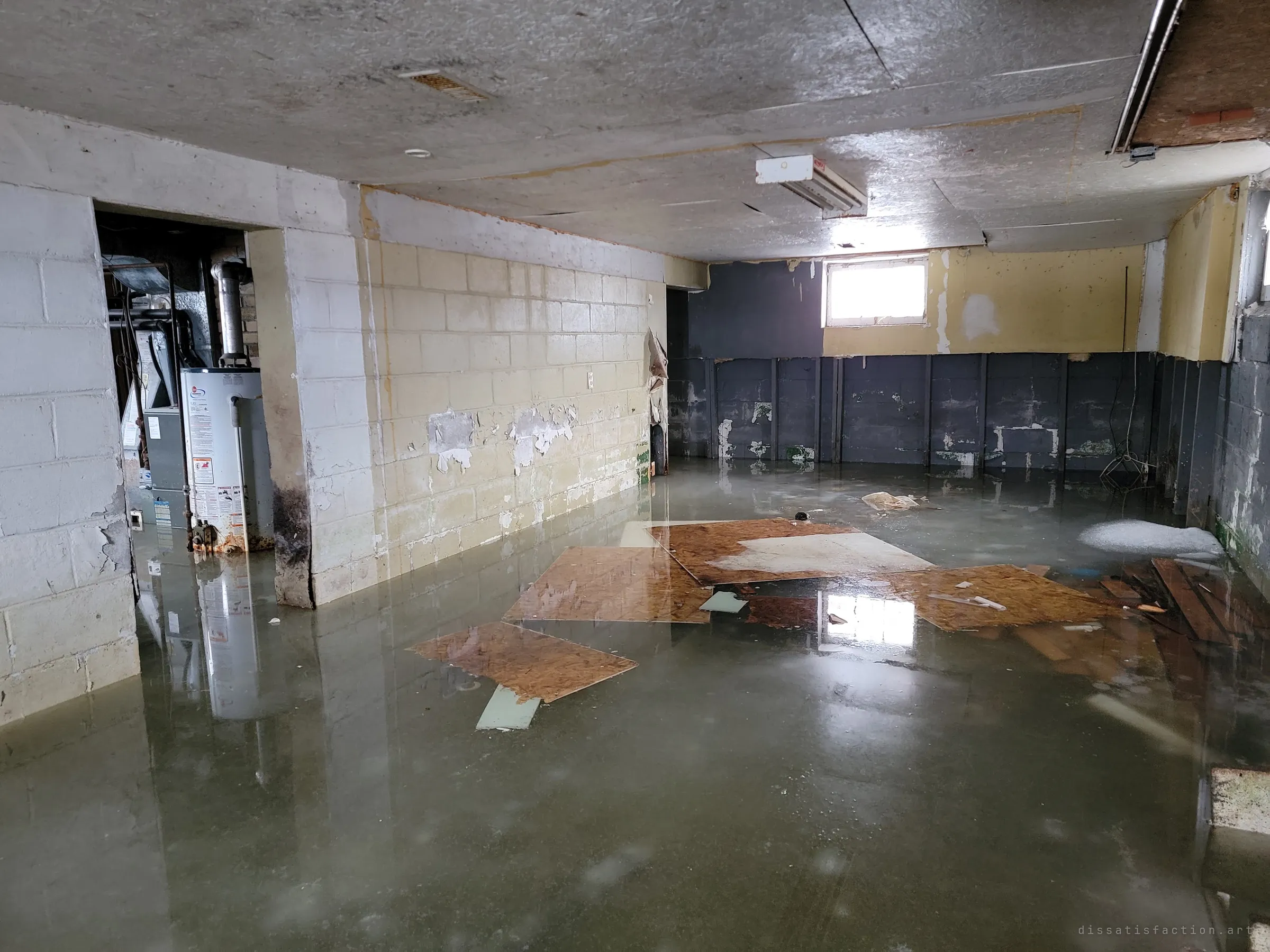 A basement flooded ankle-deep, sheets of chipboard floating on still green water under a fluorescent tube, the furnace room glimpsed through a doorway. The high window's light mirrors across the surface.
