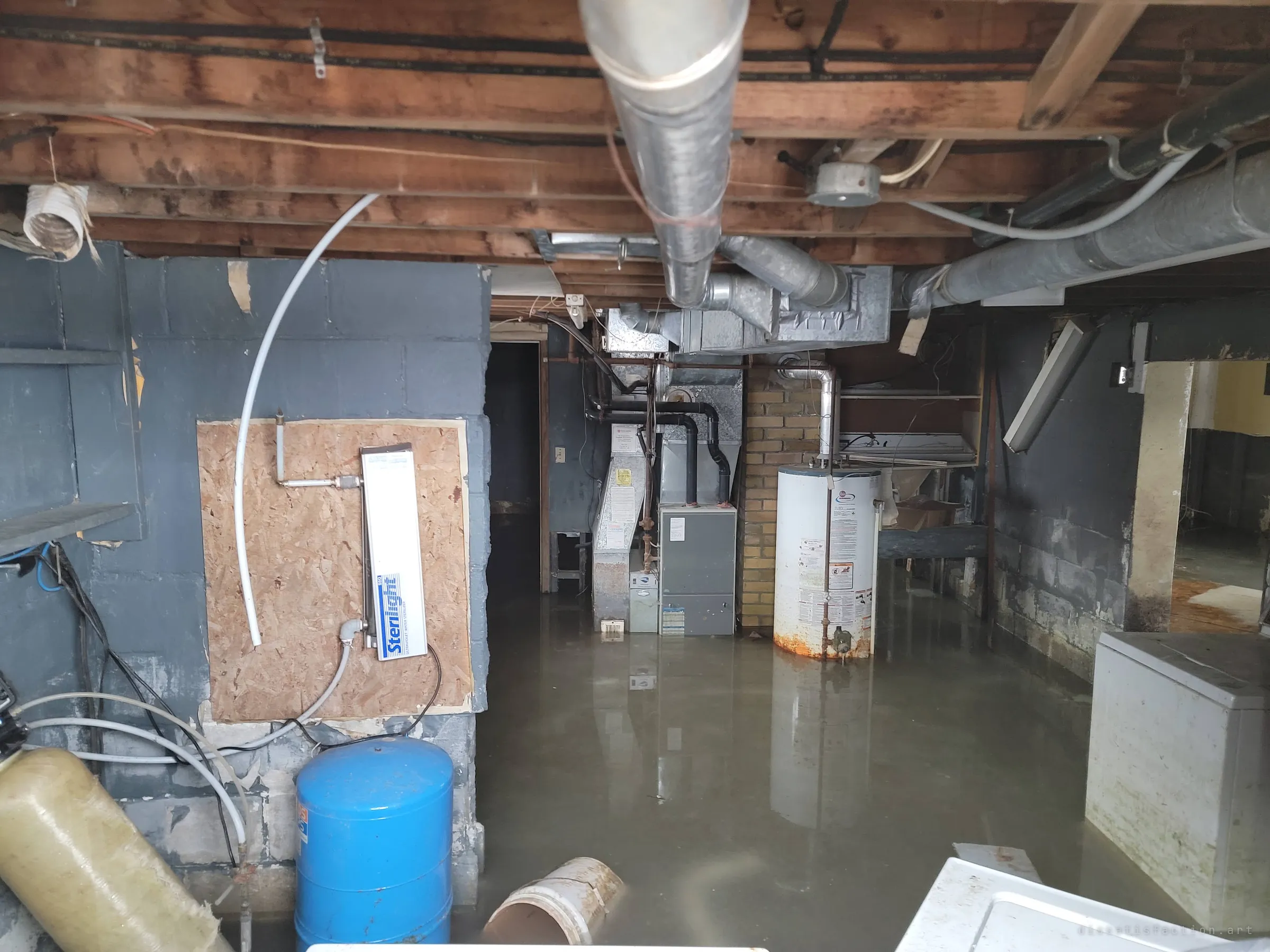The flooded utility corner under exposed joists: a rusting water heater, furnace and ducts standing in murky water, a blue pressure tank and a water sterilizer mounted on plywood. The house's machinery is drowning in place.