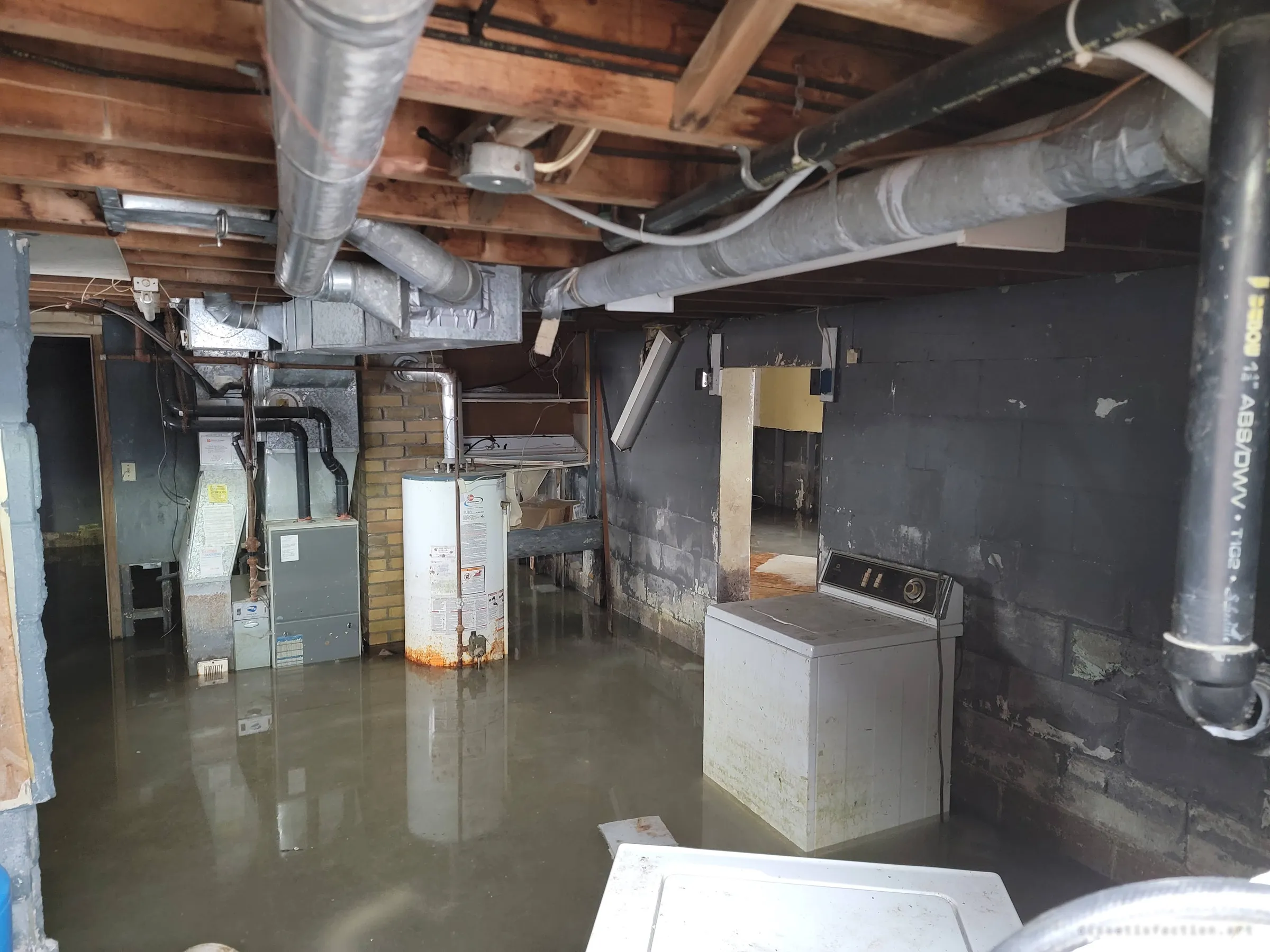 A wider view of the same flooded basement, a washing machine and dryer sitting in the water beside the furnace and rust-ringed water heater, drain pipes running down the dark block wall. Everything below the waterline is being quietly lost.
