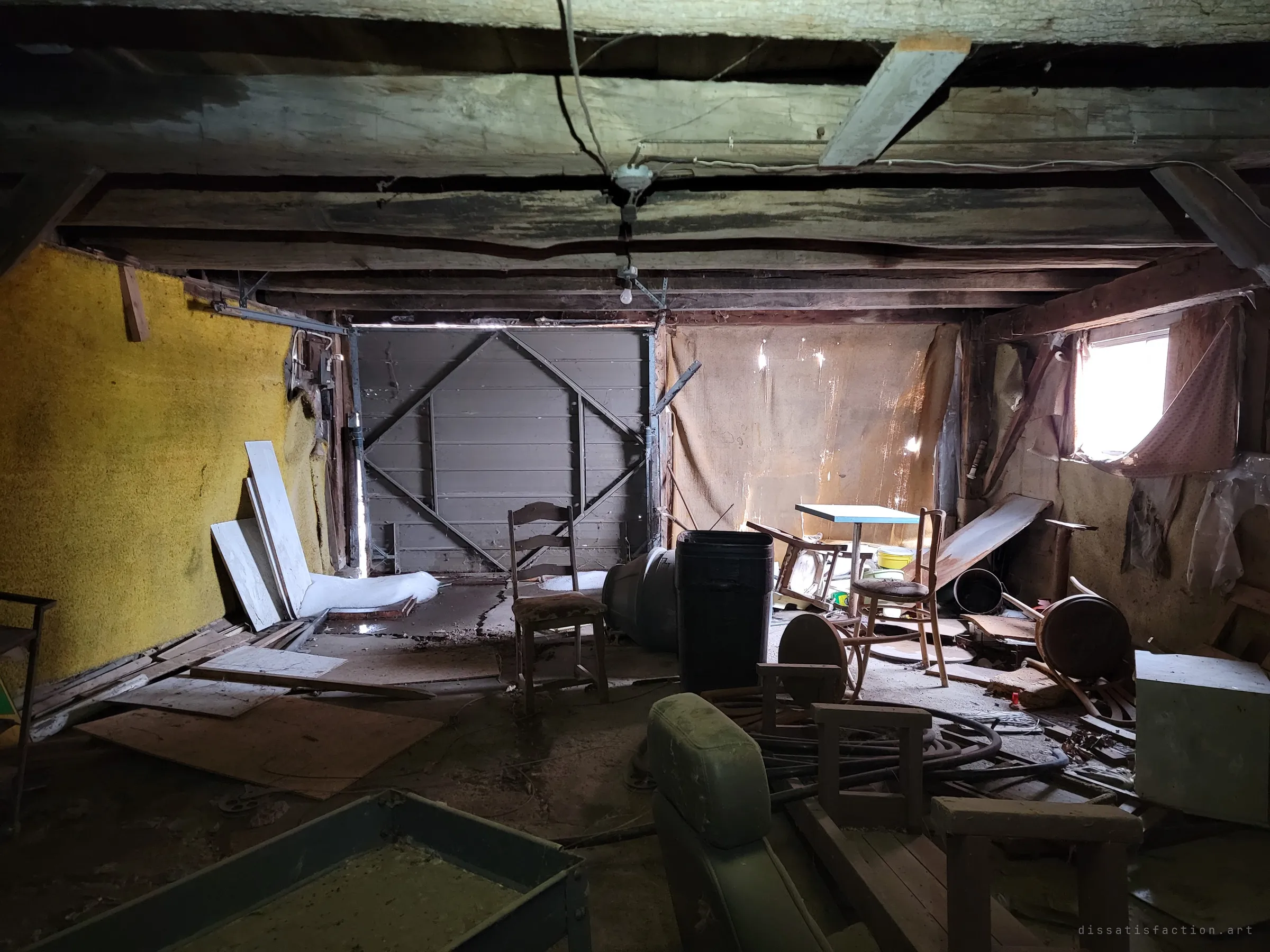 A dark garage under sagging timber beams, its panel door shut, a bare bulb hanging from a wire over scattered chairs, a small table and piles of boards. Light leaks in through a torn curtain and gaps in the walls.