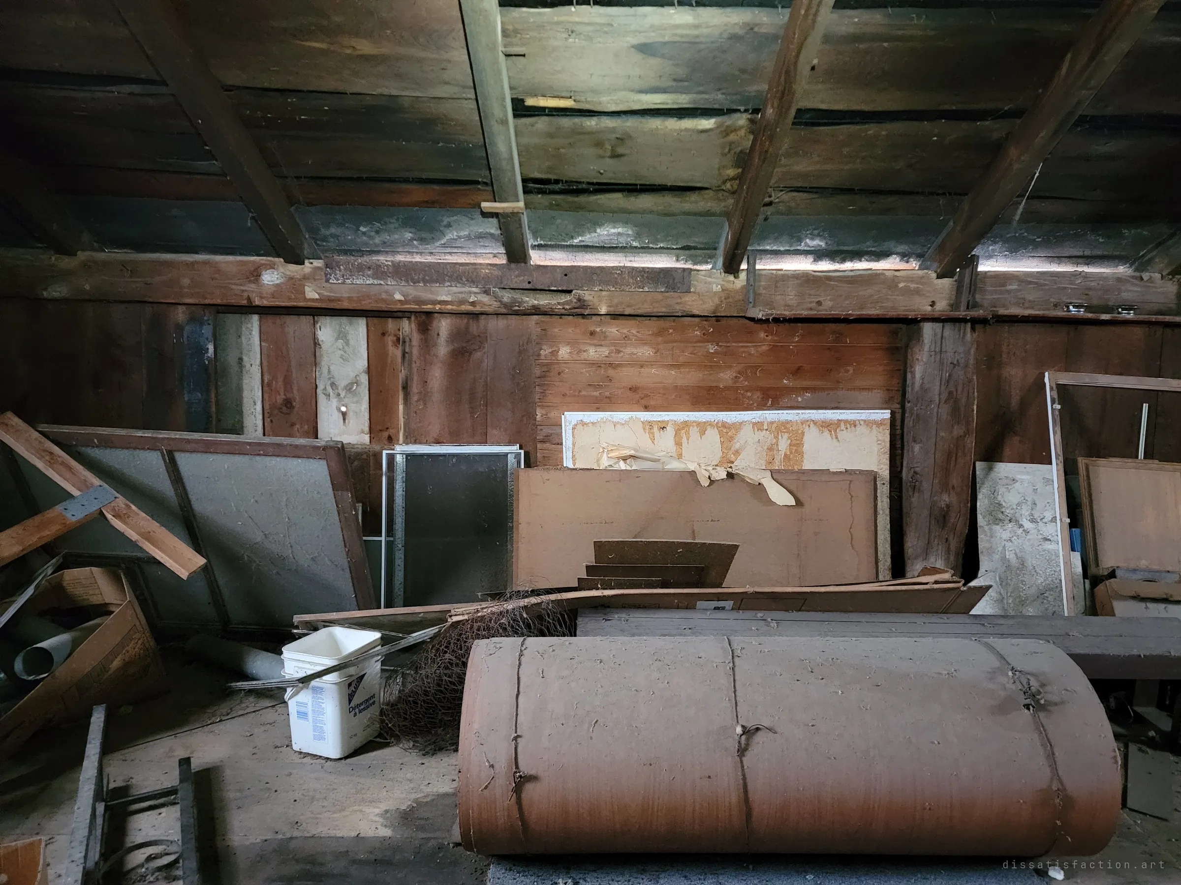 A loft of rough plank walls and rafters, old window screens, boards and a detergent bucket stacked against the wall behind a large rolled bundle of brown felt. Storage nobody came back for.