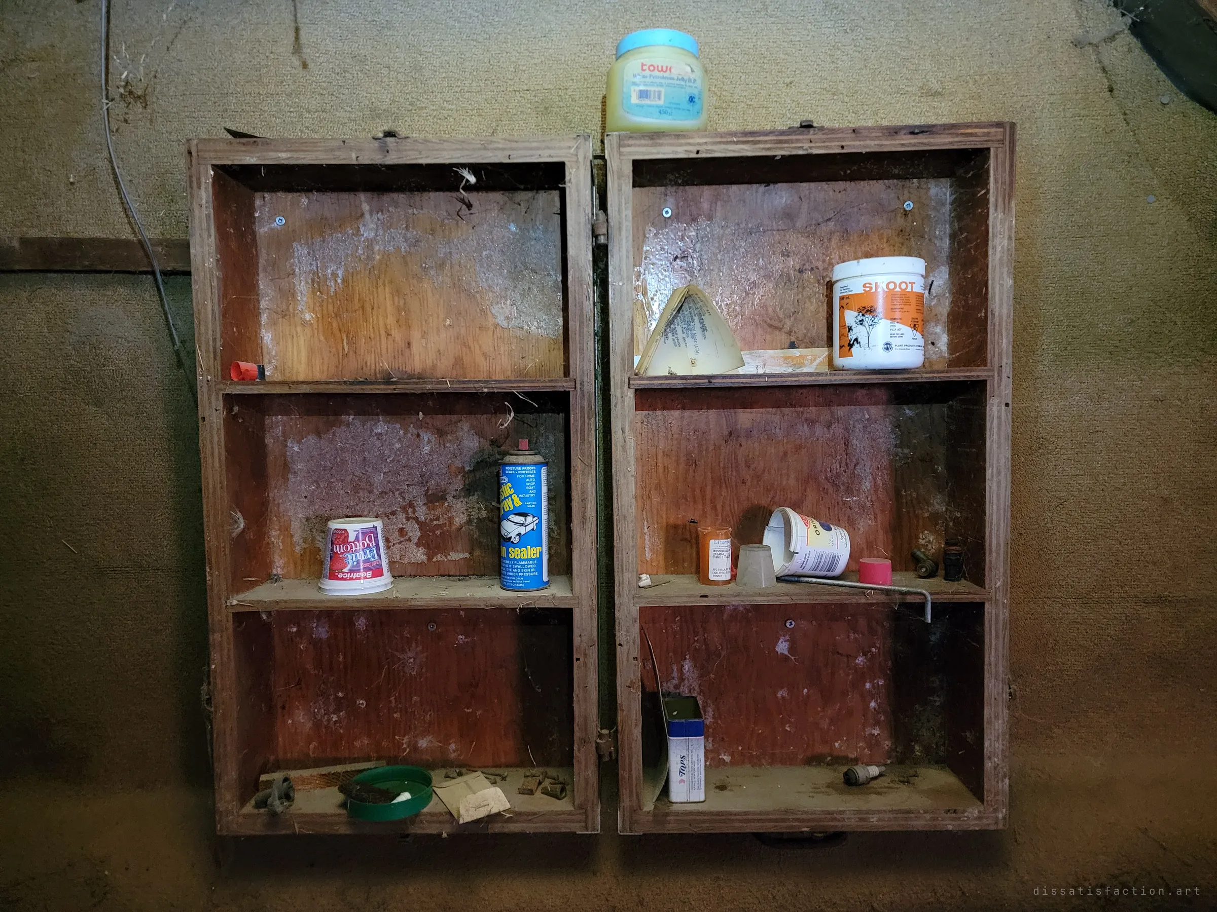 A wall-mounted wooden cabinet hangs open against a stained wall, its shelves holding a few leftover jars, a spray can, a yogurt cup and an old pill bottle. The small household leftovers show how suddenly the place was left.