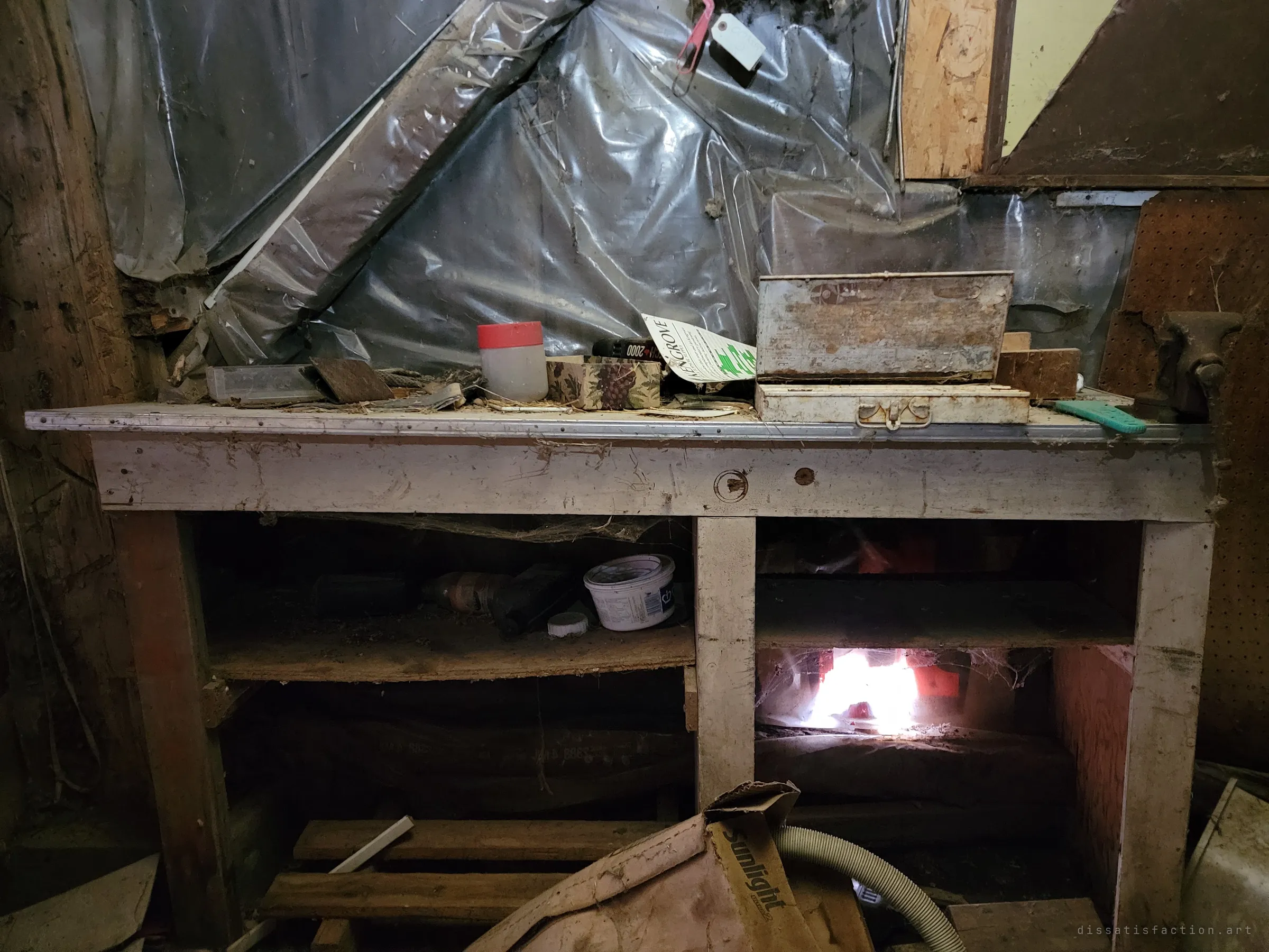A battered white workbench sits against plastic-sheeted walls, its top cluttered with tins, boxes and a bench vice while a flashlight glows from the shelf below. A working corner that was simply walked away from.