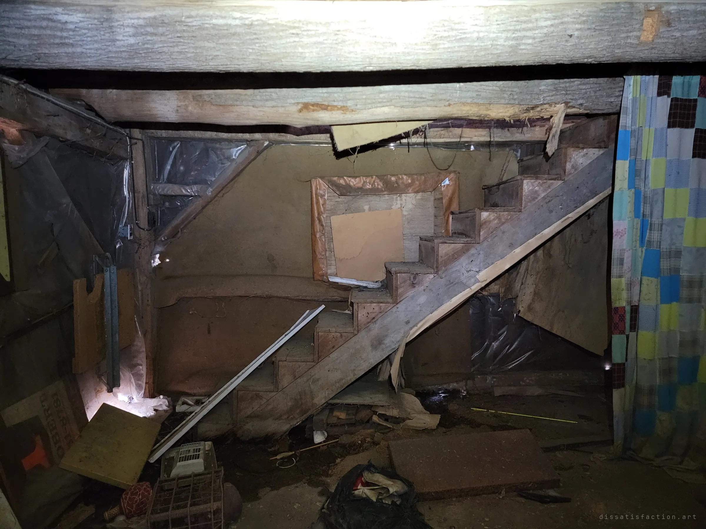 A narrow wooden staircase climbs through a low, cramped basement under heavy timber beams, with a patchwork quilt hung as a curtain on the right. Debris and loose boards cover the dirt floor.