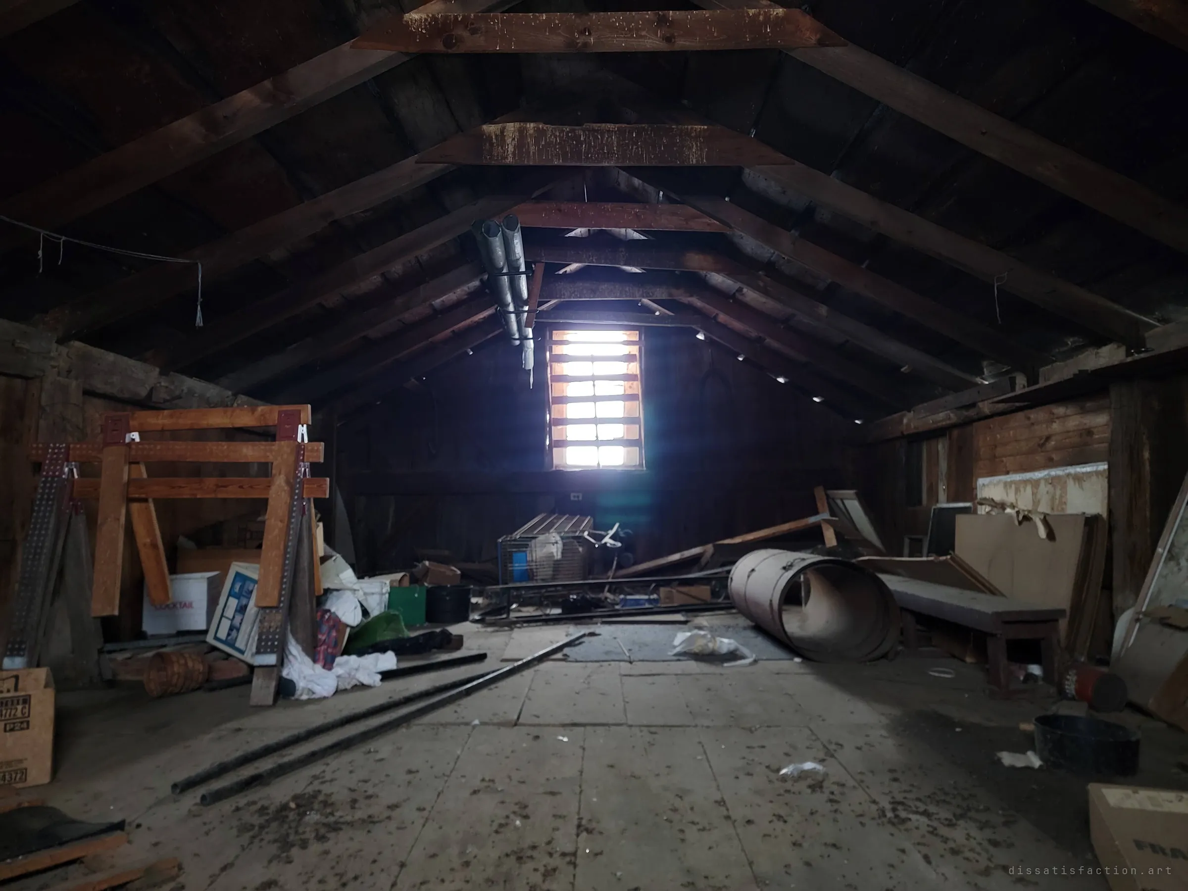 A long timber-framed attic stretches toward a single slatted gable window that throws a cold beam of light across the dusty floorboards. Sawhorses, boxes and a rolled metal drum are scattered along the sides.