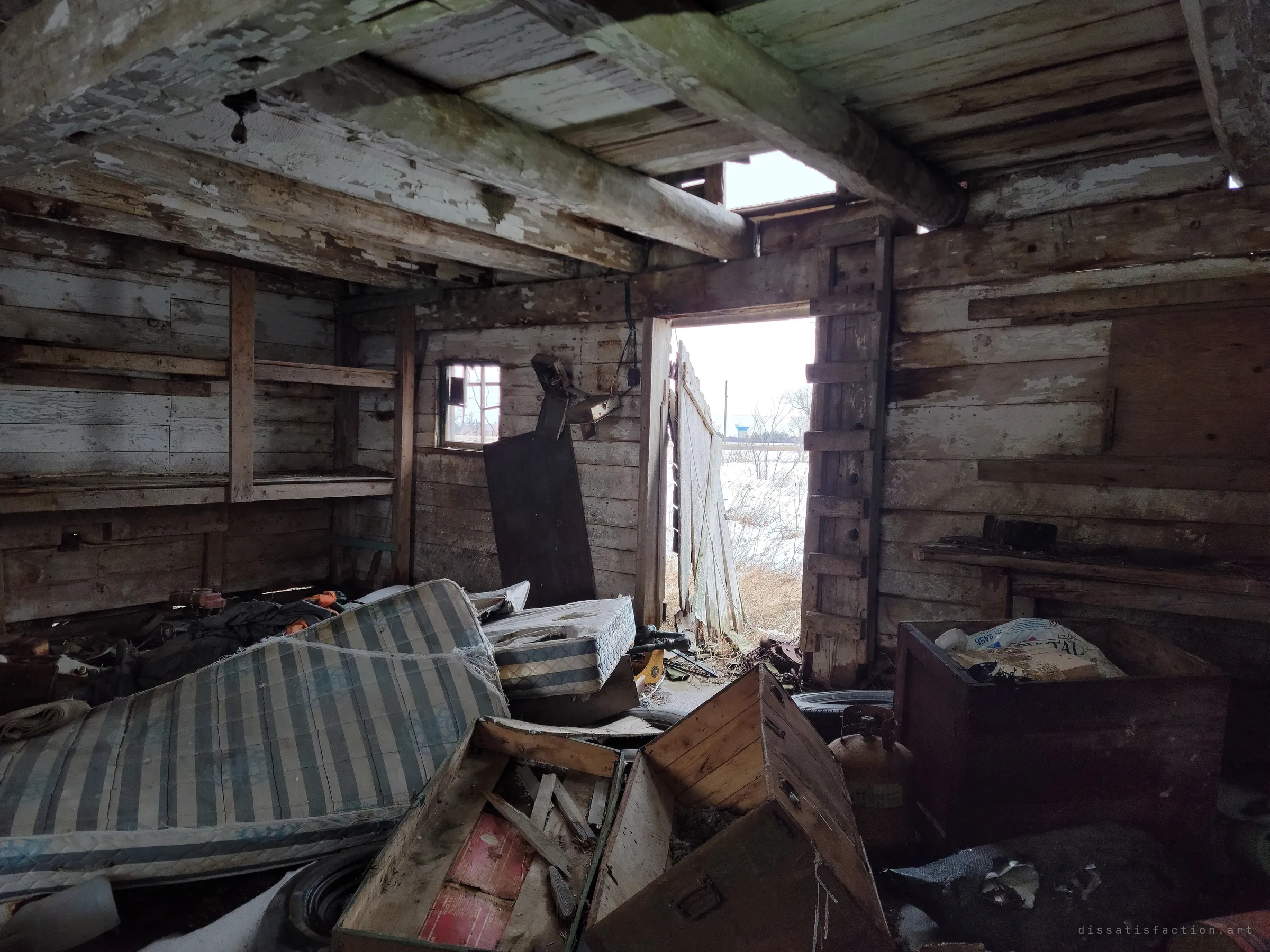 Inside a weathered log outbuilding, collapsed mattresses and broken crates fill the floor while a sagging door hangs open onto a snowy field. Peeling whitewash and daylight through the roof show how far the building has decayed.
