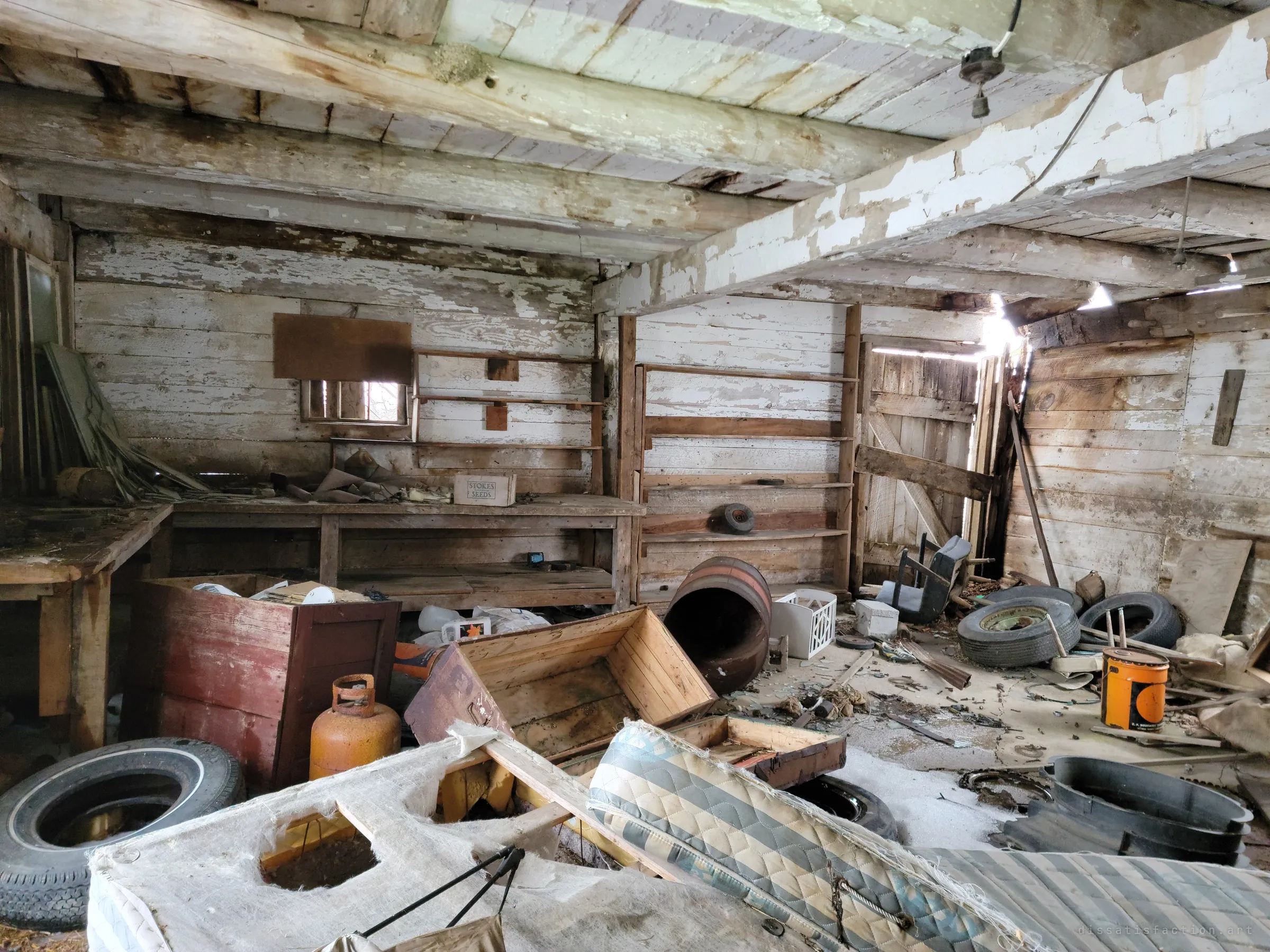 A wide view of the whitewashed log workshop shows empty shelving, an old workbench, overturned crates, tires and a propane tank strewn across the floor. Light leaks in through gaps in the far wall and roof.