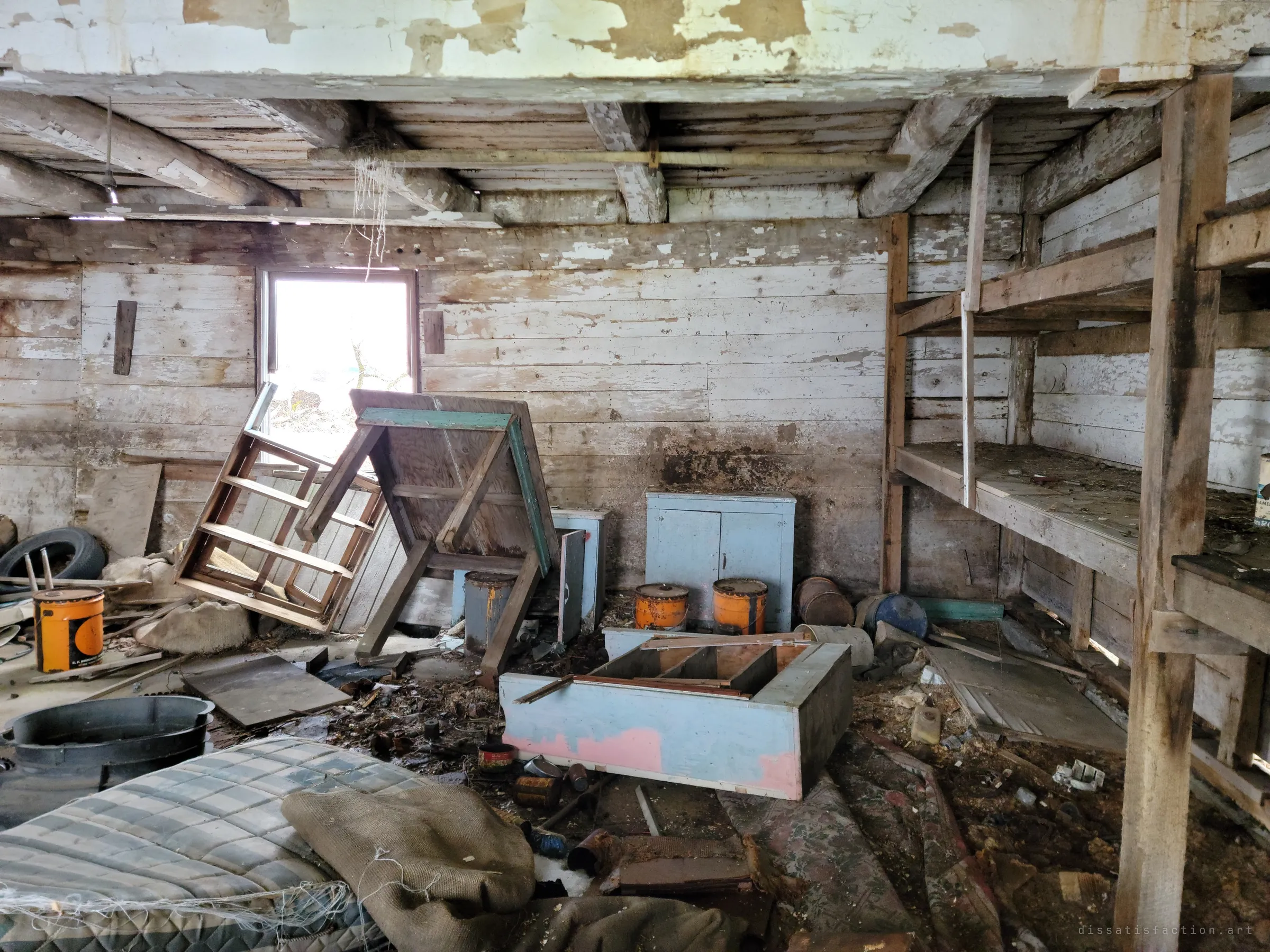 Toppled chairs and a pastel cabinet lie in the rubble beneath a bright window, with rough wooden shelving along the right wall. Paint cans and old mattresses fill the rest of the room.