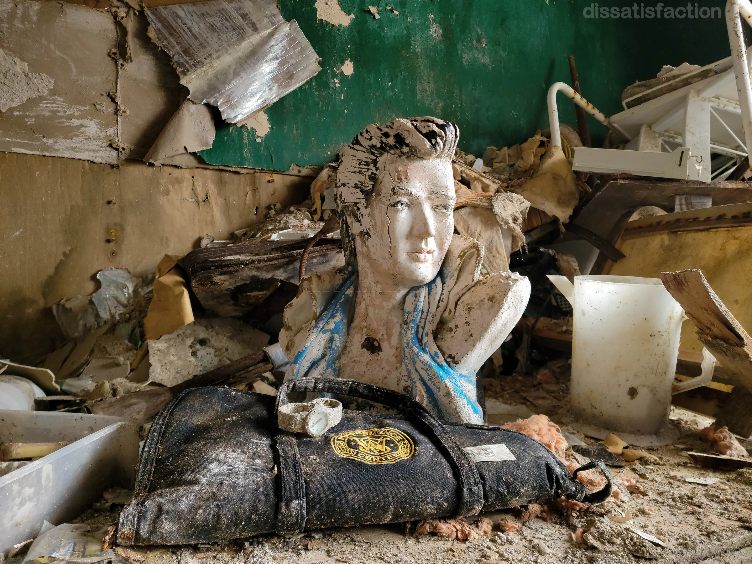 A cracked plaster bust with slicked-back hair sits in the debris against a peeling green wall, a mud-caked wristwatch resting on a dark carry bag in front of it. A strange, almost shrine-like arrangement left behind in the ruin.