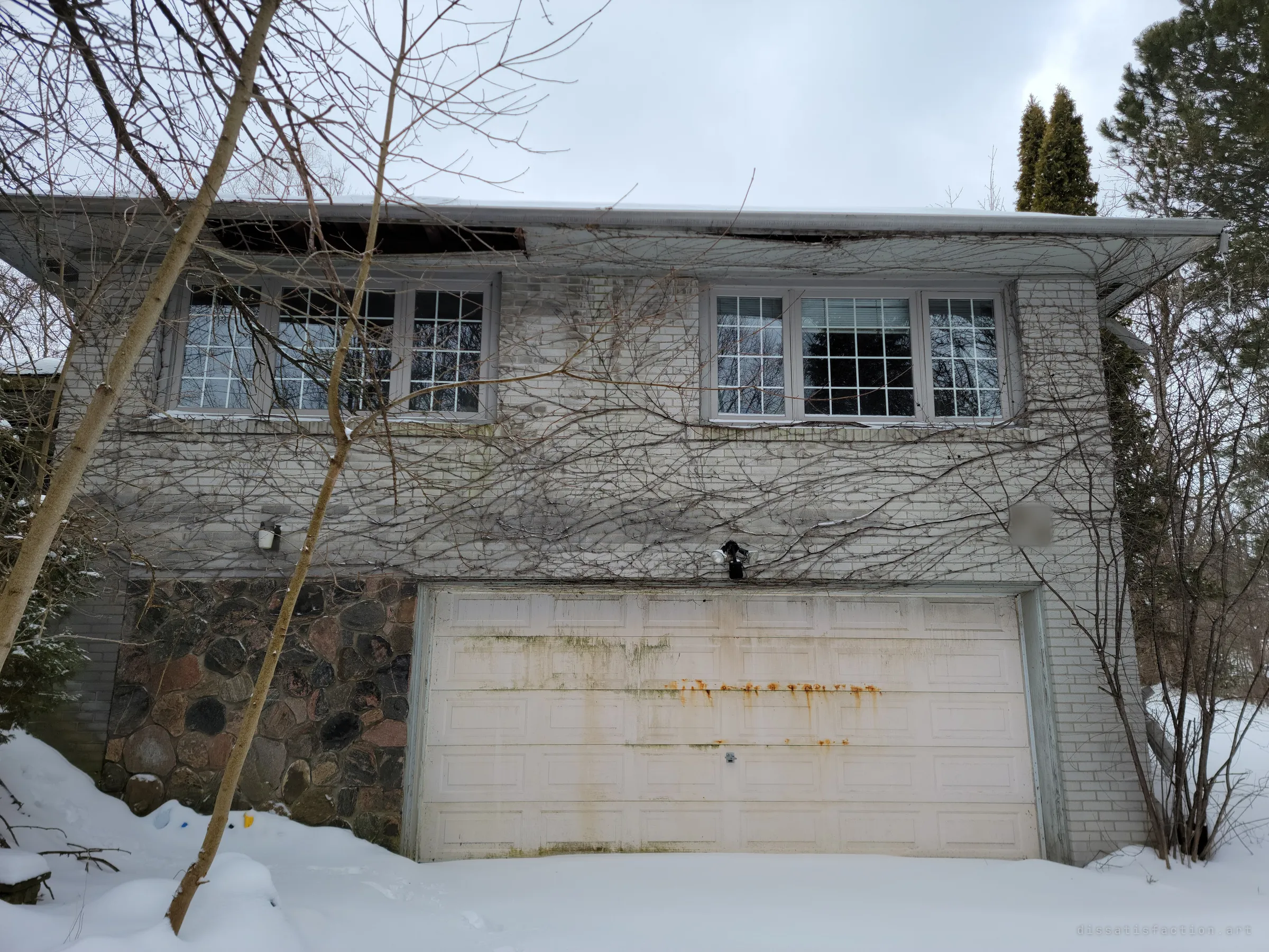 The front of an abandoned split-level house in winter, bare vines crawling over pale brick above a rust-streaked garage door and snowdrifts. The collapsing soffit hints at the damage inside.