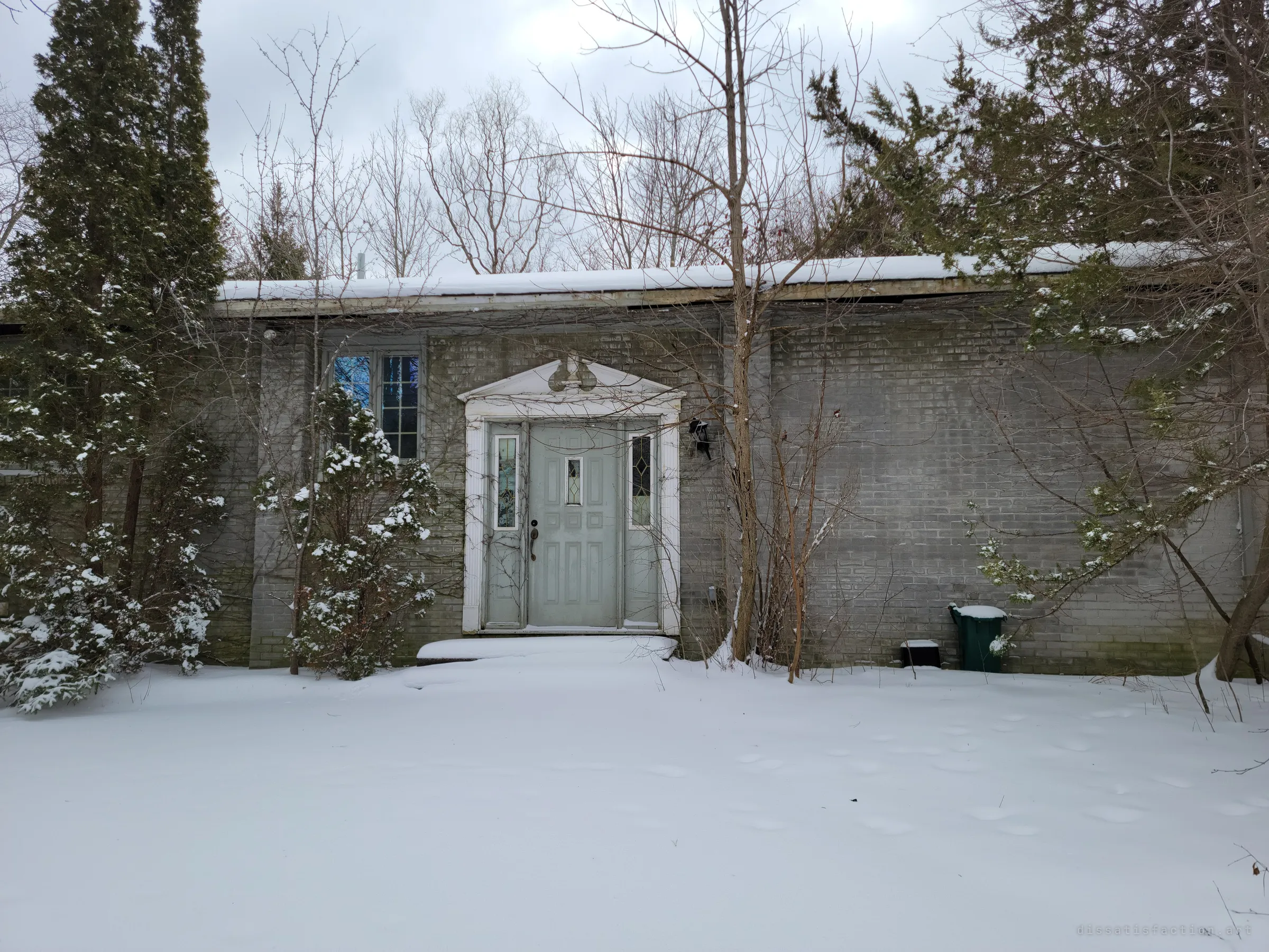 A pale green front door with a white pediment, half hidden by snowy evergreens and bare saplings in an untouched snowy yard. The quiet approach to the abandoned house.
