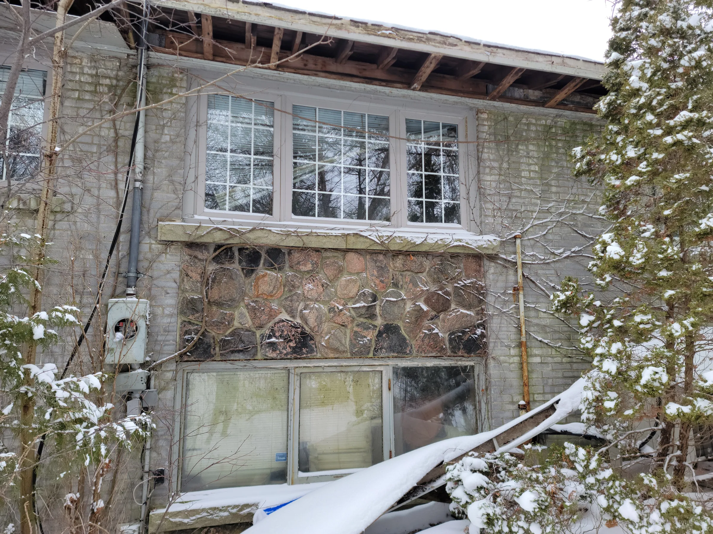 Looking up at the back wall of the house: exposed rafters where the soffit has fallen away, gridded windows over a fieldstone band and a fallen gutter half buried in snow. The structure is visibly coming apart.
