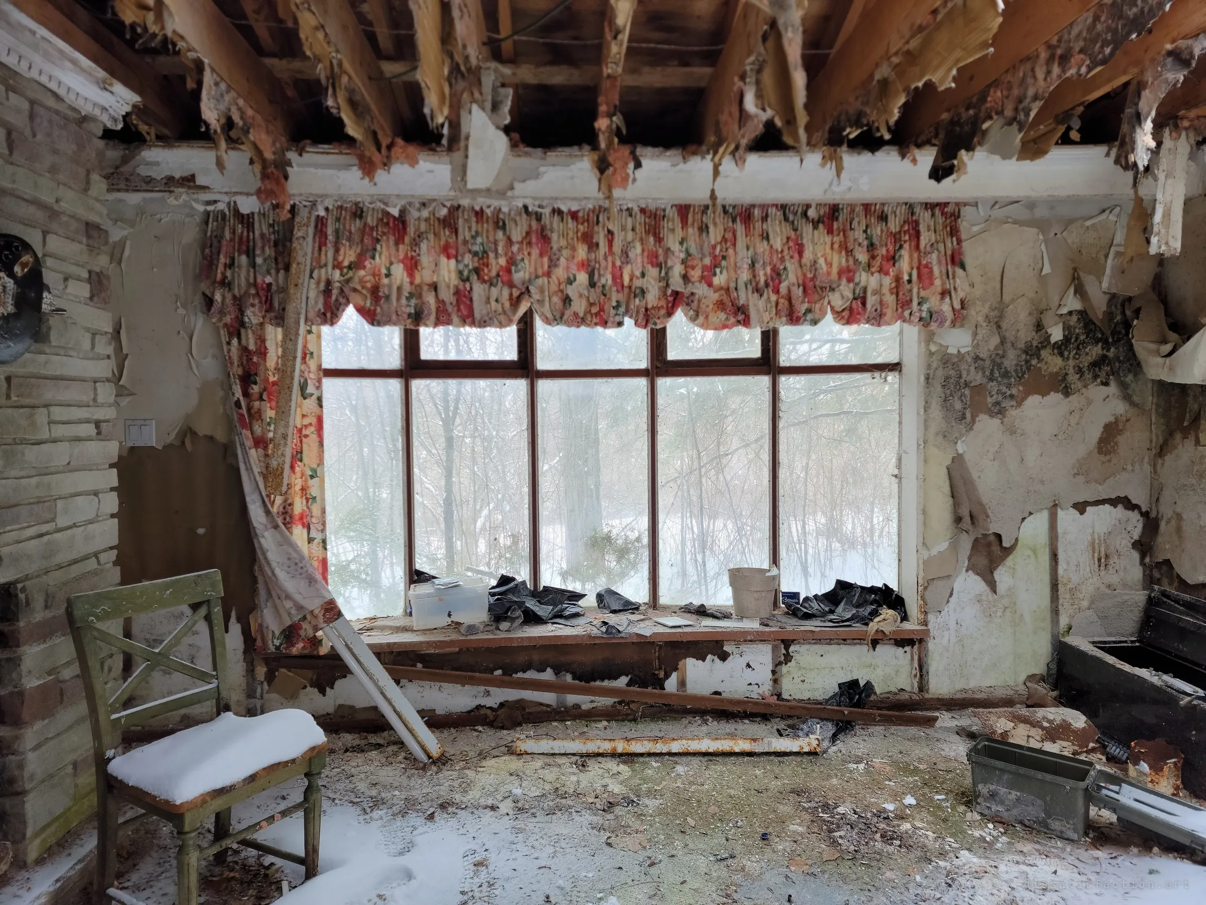 A living room open to the joists, floral valance still hanging over a wide window onto snowy woods, with snow drifted onto a lone chair and the floor. The roof has failed enough to let winter inside.