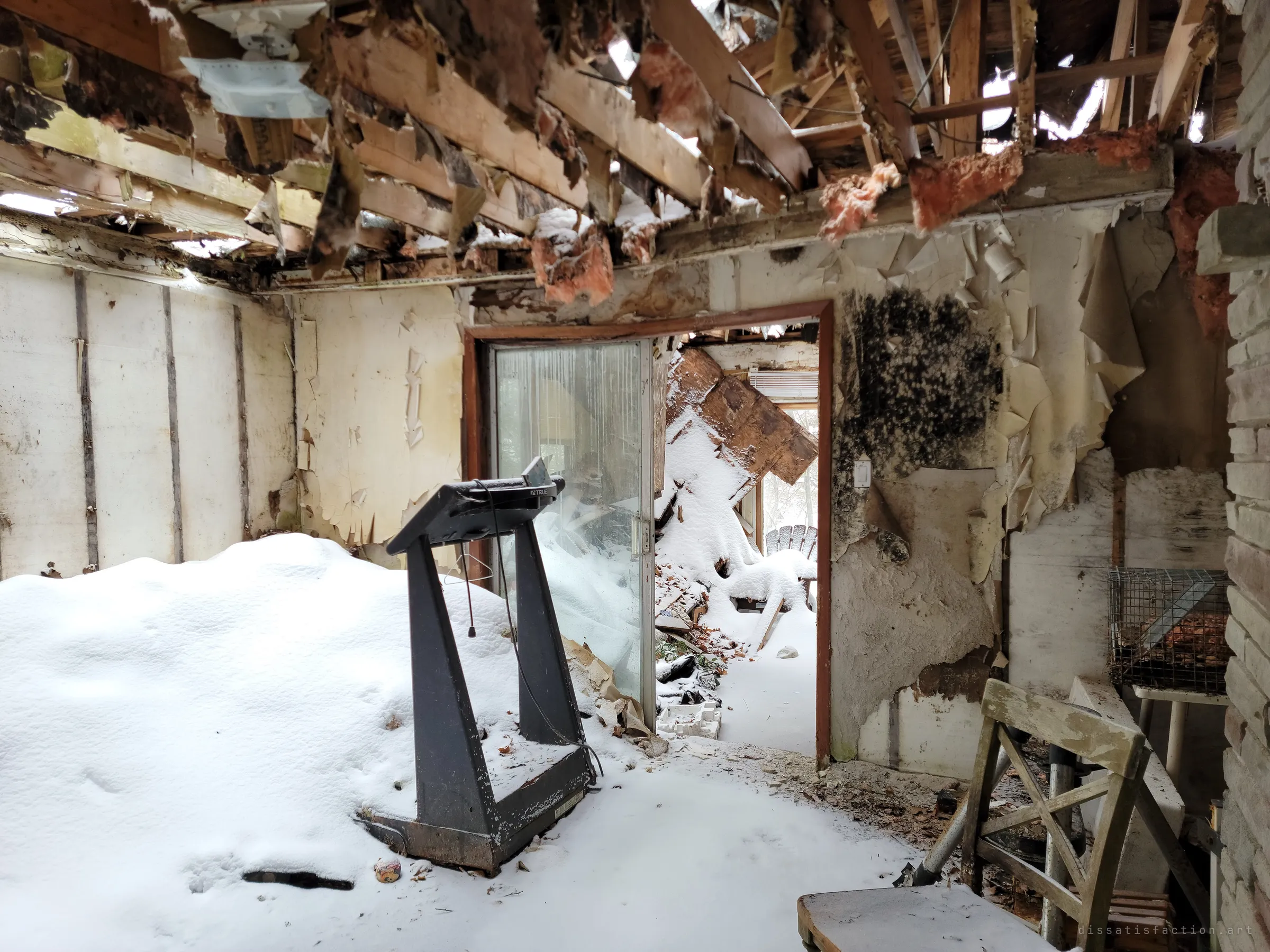 An old treadmill stands in a room buried under a snowdrift, the ceiling torn open to the rafters and black mould spreading across the wall by a sliding door. Nature is reclaiming the house from above.