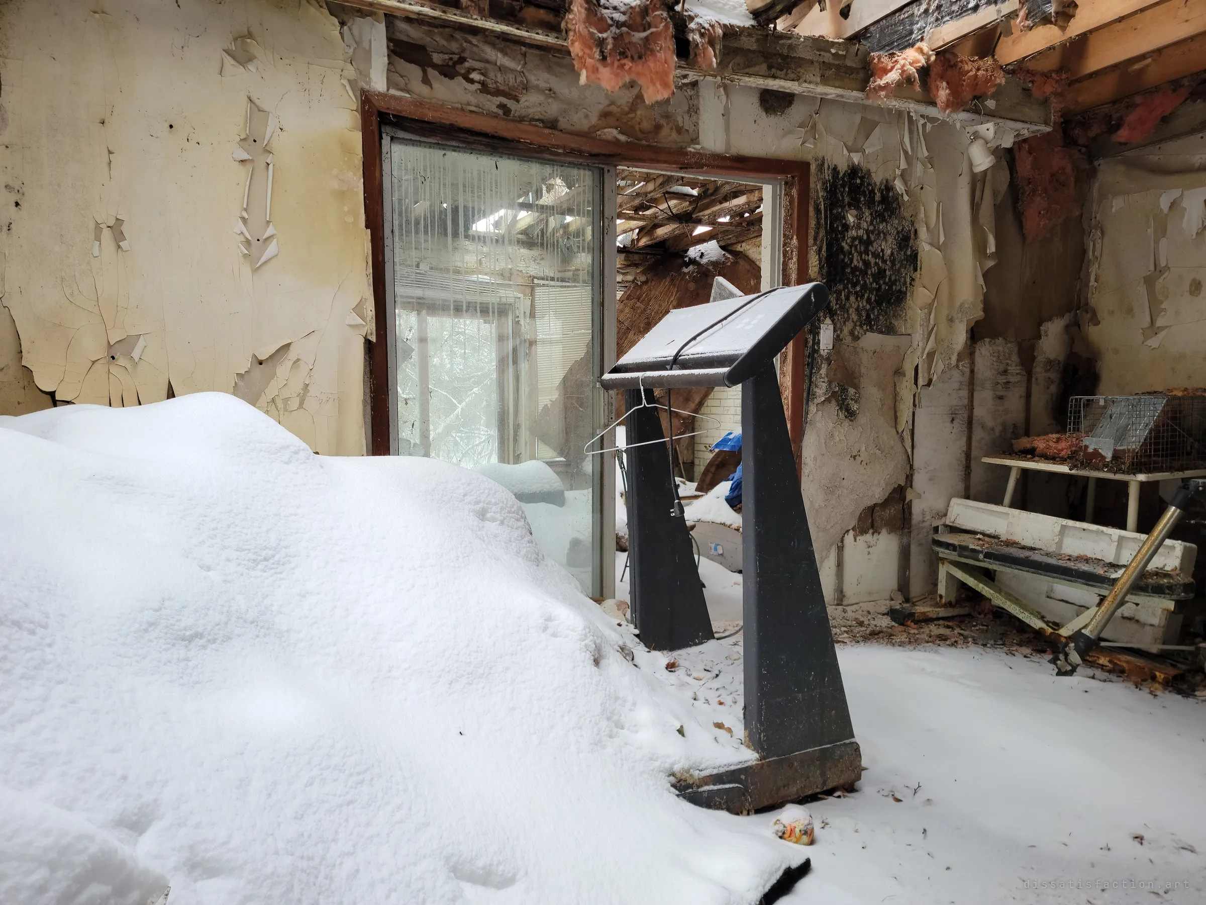 A closer view of the snow-covered treadmill with a wire hanger dangling from its console, beside a mouldy sliding door and an animal trap on a table. The same room seen low and near.