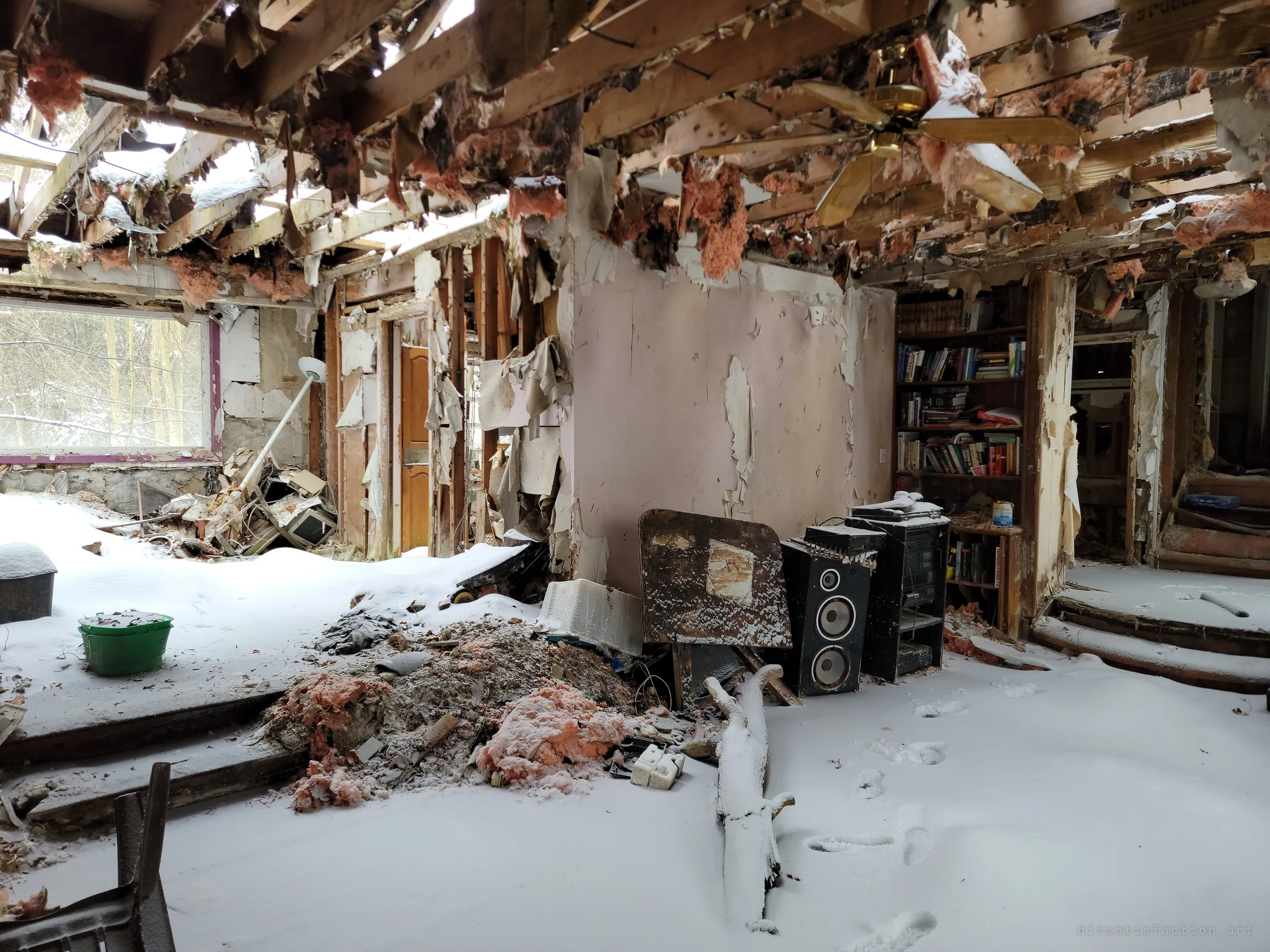 A snow-filled living room under a collapsed ceiling, with a brass ceiling fan, a stereo and speakers, and a bookcase still full of books. A whole household left to the weather.