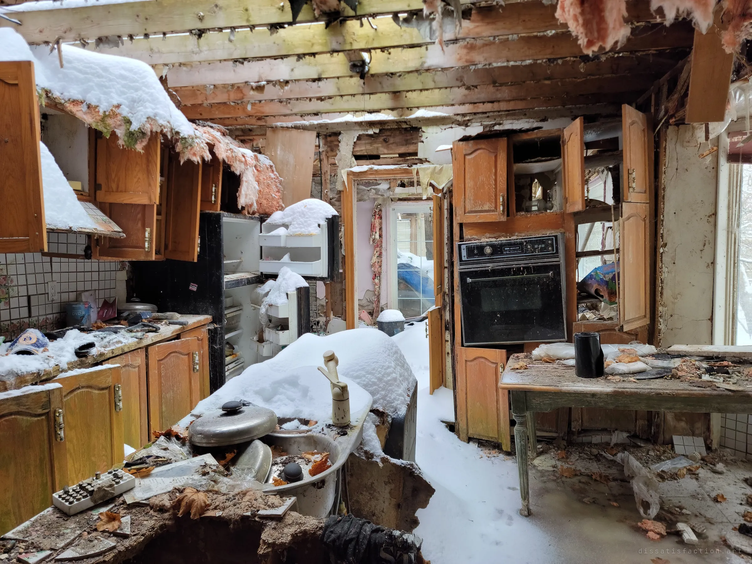 A snow-filled kitchen under a caved-in ceiling, oak cabinets hanging open over a sink and wall oven buried in drifts. The roof is gone, so winter has moved straight into the room.