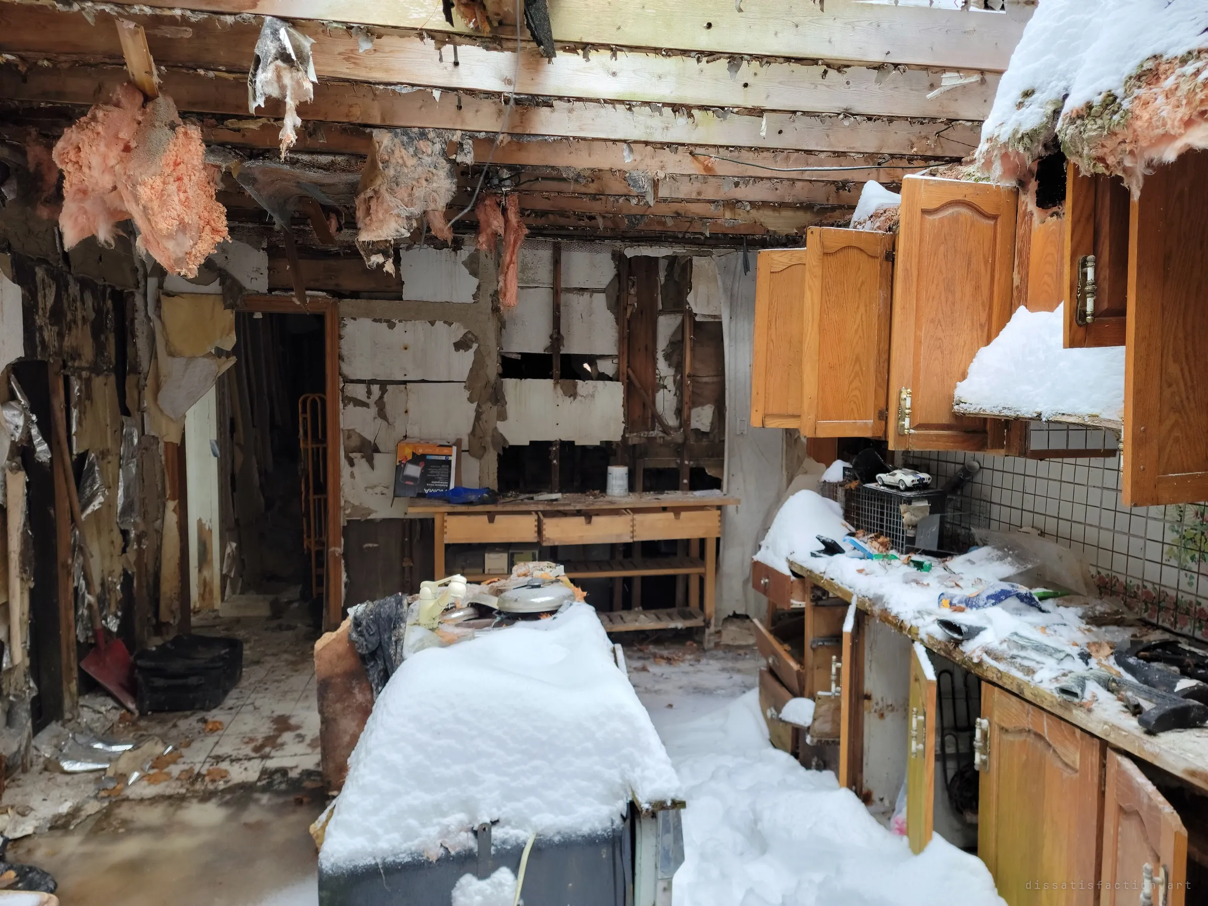 The same kitchen looking the other way: pink insulation dangles from bare joists above a snow-covered island and a counter strewn with tools and a toy car. Stripped walls show the house gutted down to its studs.