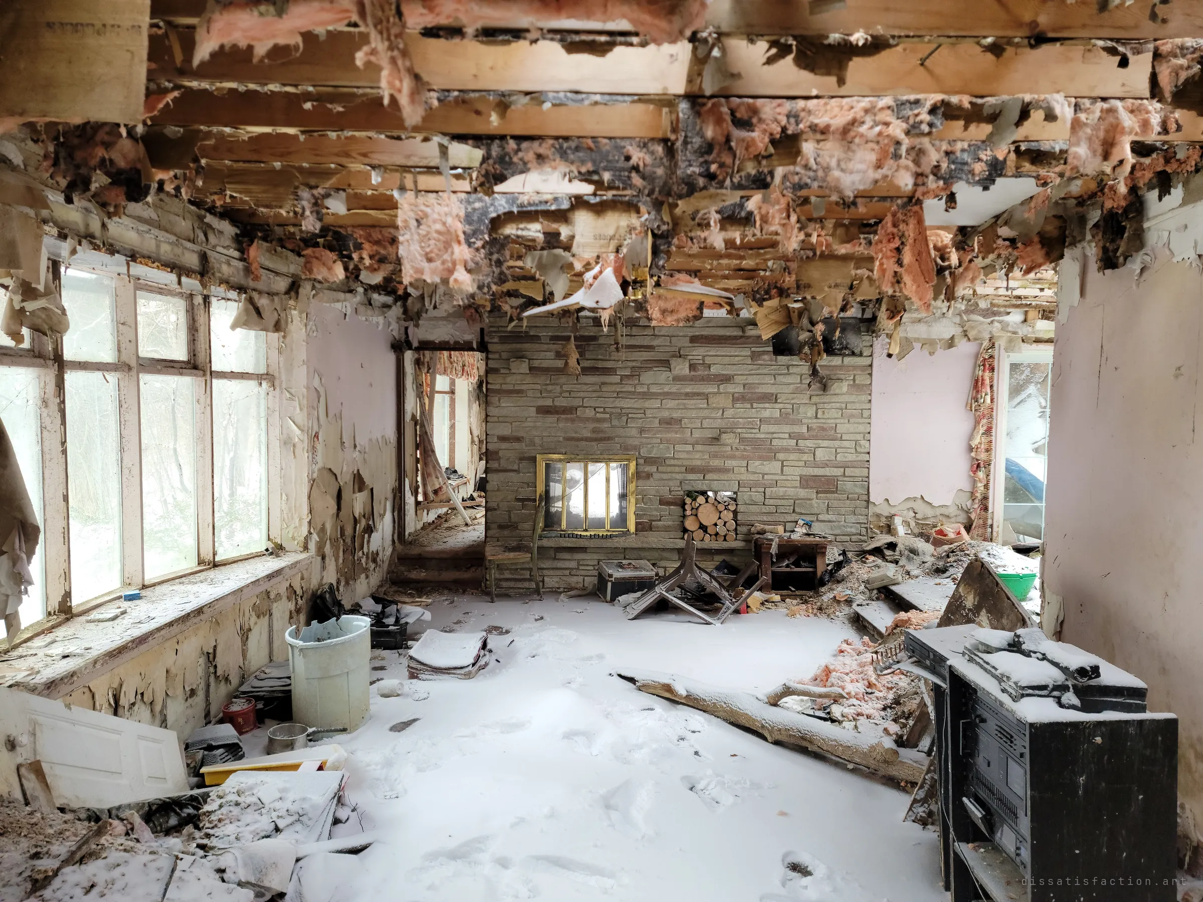 A living room with a stone fireplace wall, its ceiling torn open and shedding insulation onto a snow-dusted floor full of footprints and toppled furniture. A stereo cabinet still stands in the foreground.