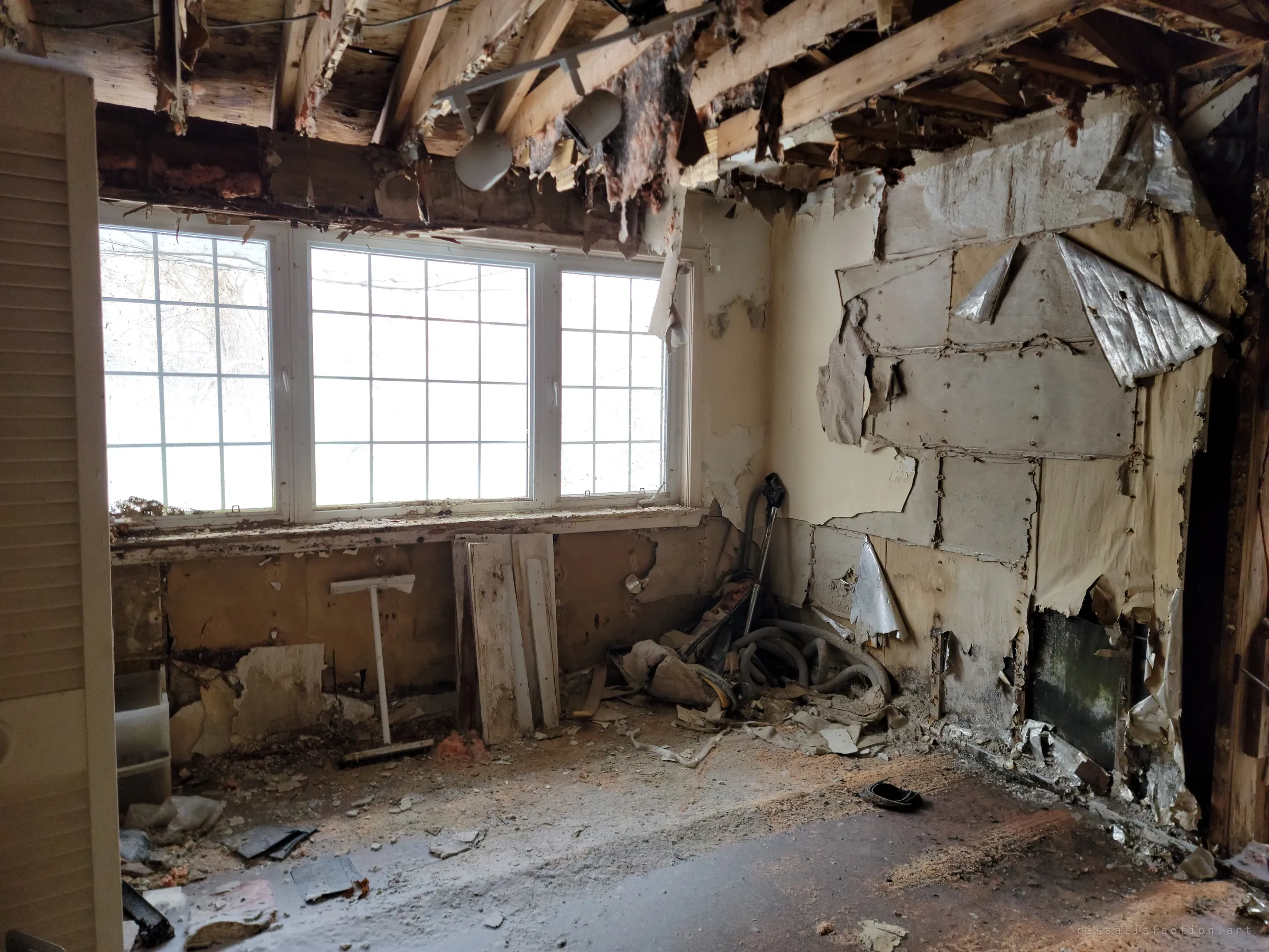 A bright bedroom corner with a triple window, peeled wallboard and foil vapour barrier curling off the walls. A vacuum hose and broken boards are piled beneath the sill.
