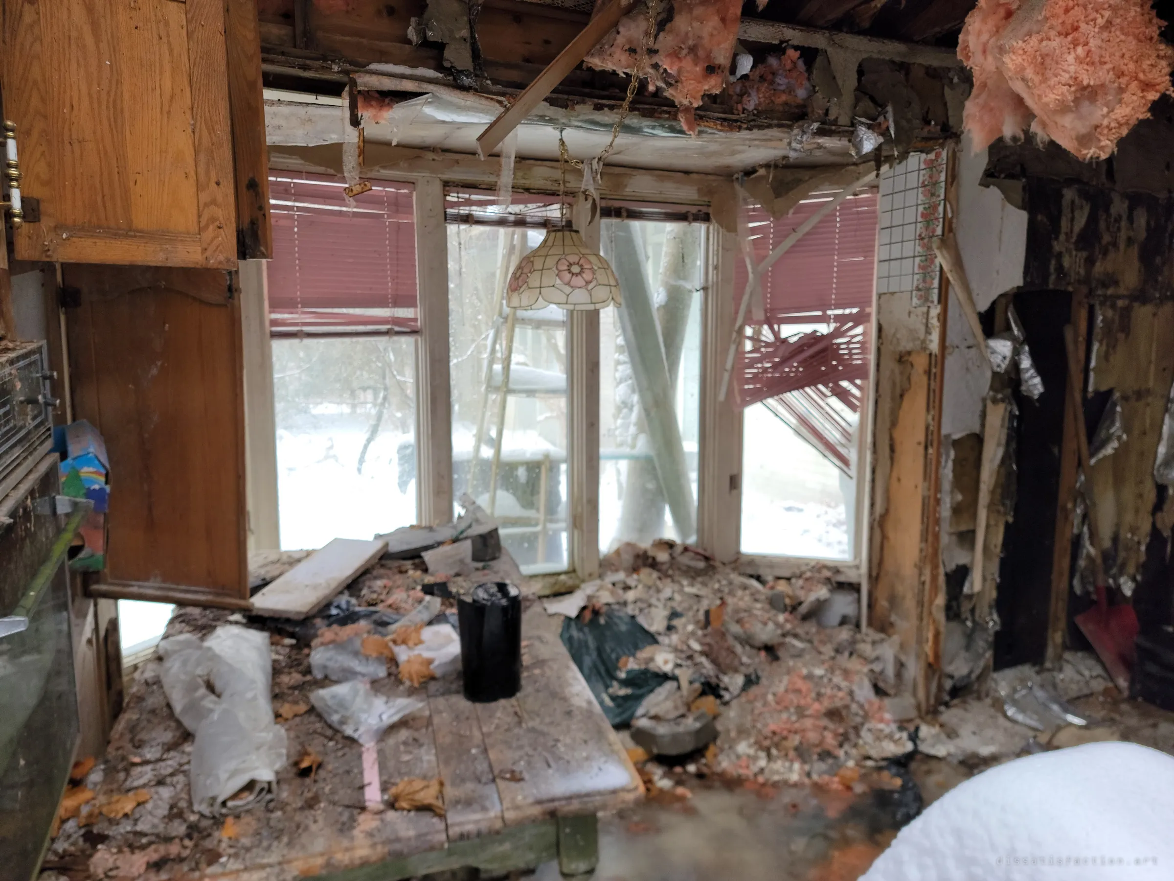 A breakfast nook under a flowered stained-glass lamp, the table buried in plaster debris and dead leaves. Pink blinds hang torn across windows looking out on snowy trees.