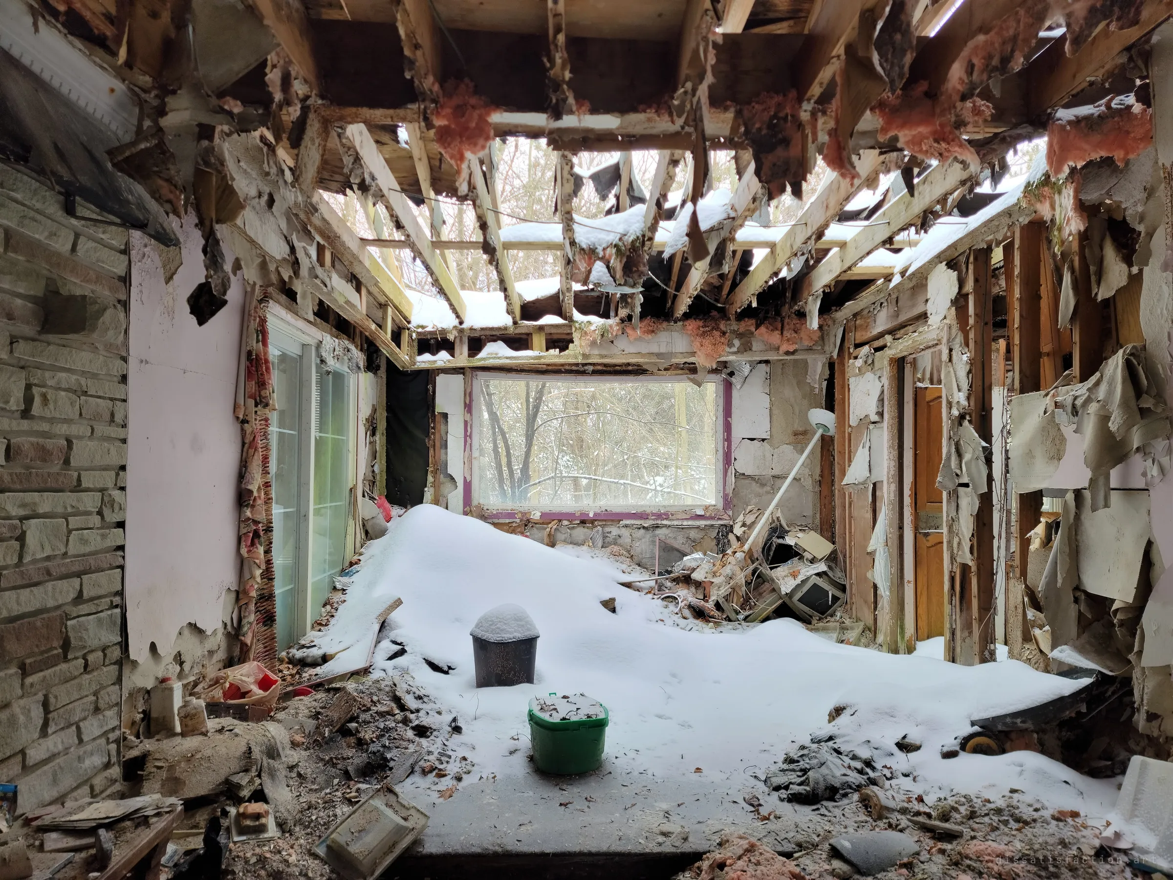 A room open to the sky, rafters laced with snow and a deep drift sloping across the floor toward a window of bare winter trees. Only a bucket and a bin stand clear of the snow.