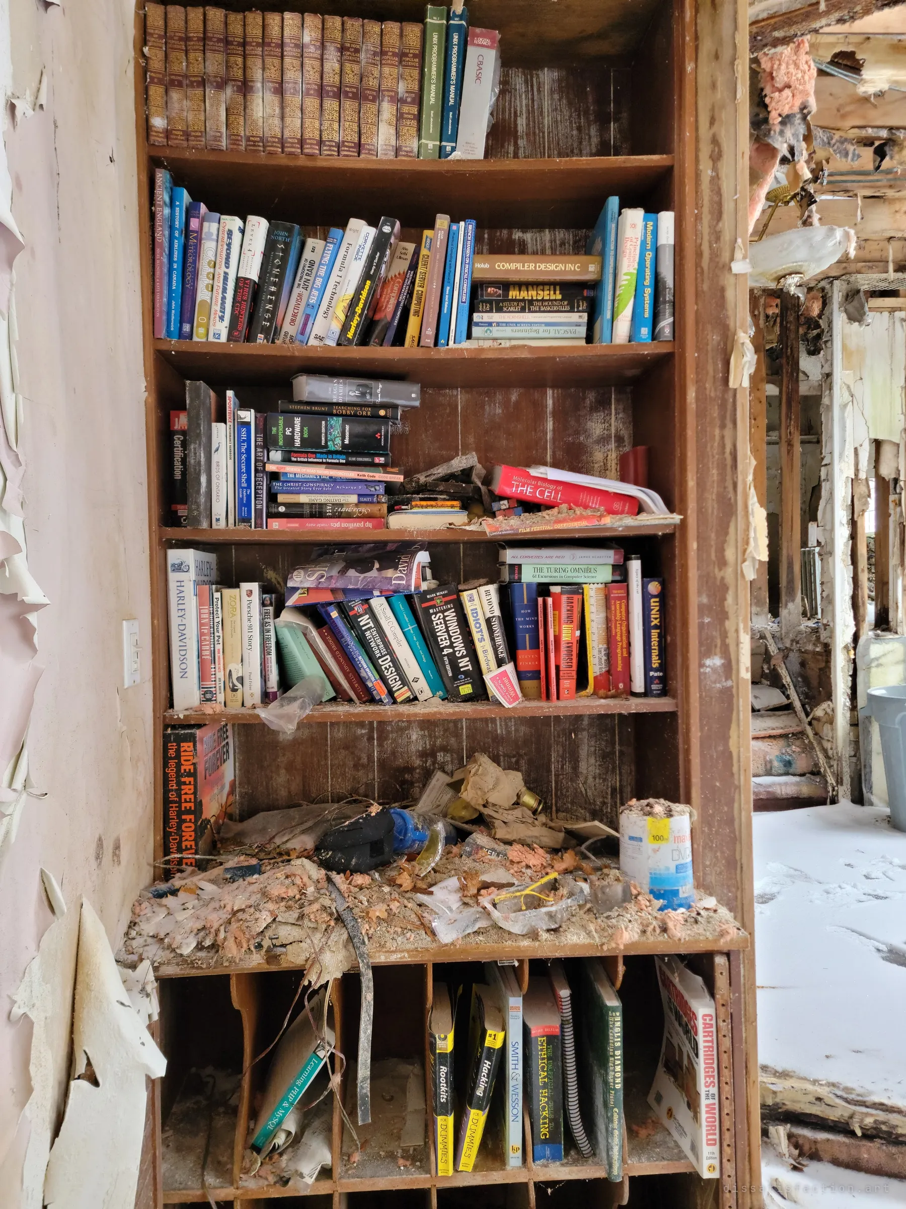 A tall built-in bookshelf still packed with programming manuals, motorbike books and old encyclopedias, the lower shelf buried in insulation. The owner's interests survive the collapse around it.