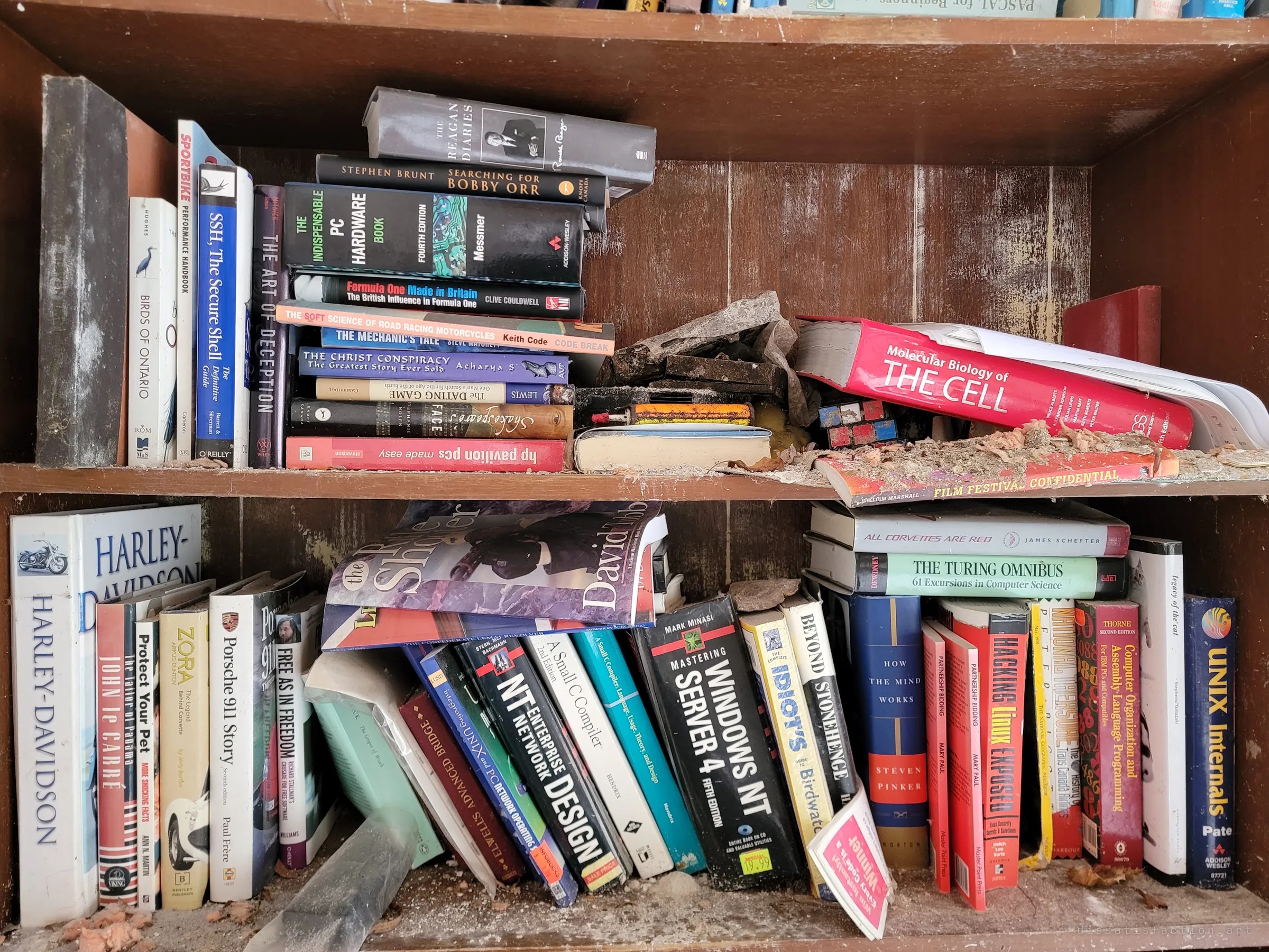 A close look at two shelves of computing, racing and motorcycle books, dusted with fallen plaster. Titles on hacking, old operating systems and Harley-Davidson sketch the person who lived here.