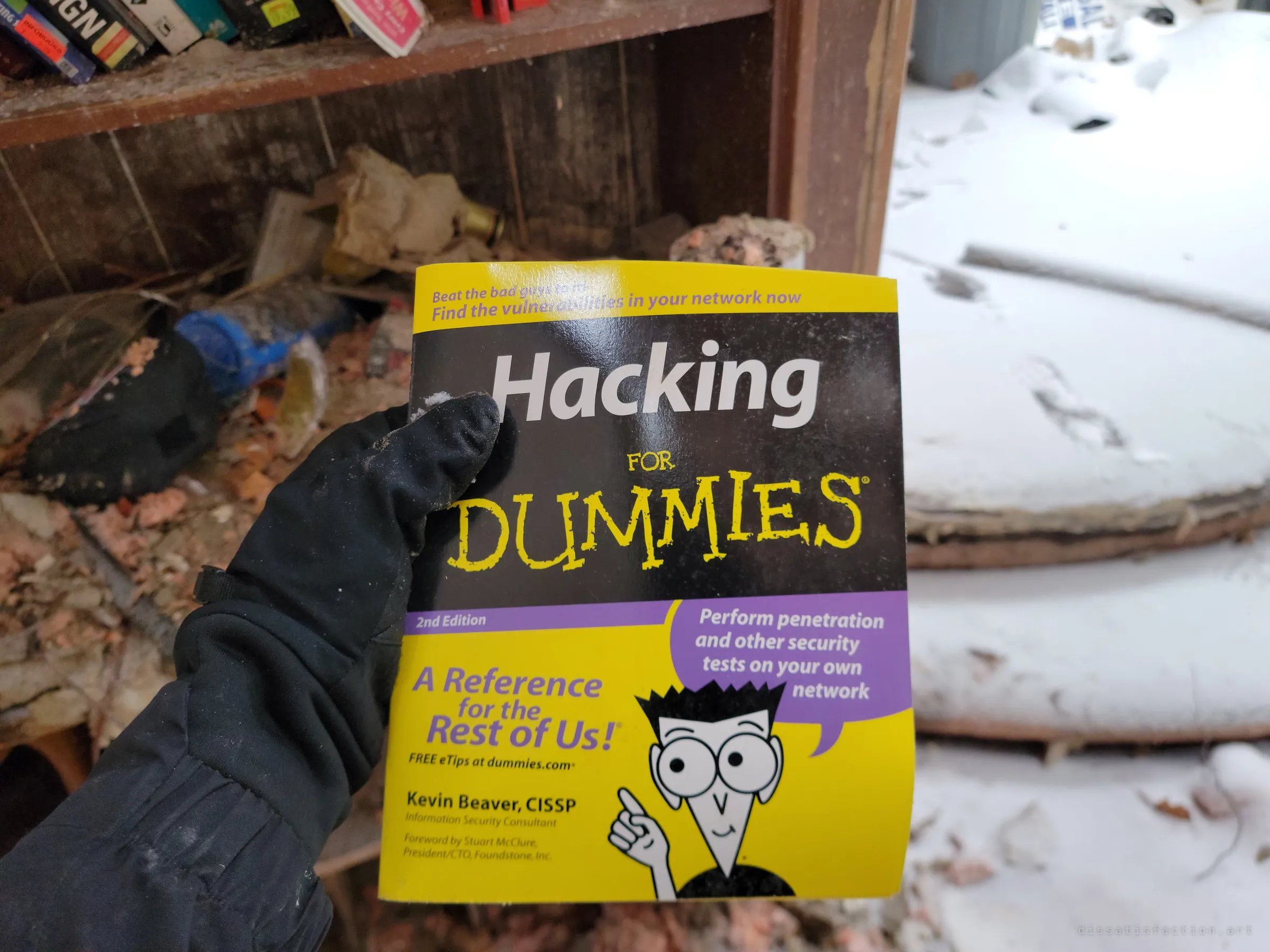 A gloved hand holds up a beginner's hacking handbook in front of a rotting bookshelf, snow drifting in through the open side of the room. One of several odd books pulled from the shelves.