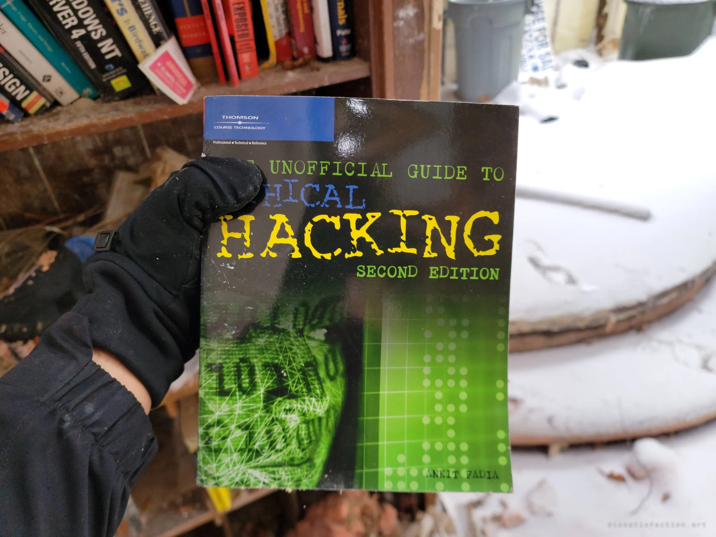 A gloved hand holds an ethical hacking guide in front of a shelf of old computer manuals, with snow piled inside the collapsing room. The book collection hints at the previous owner's interests.