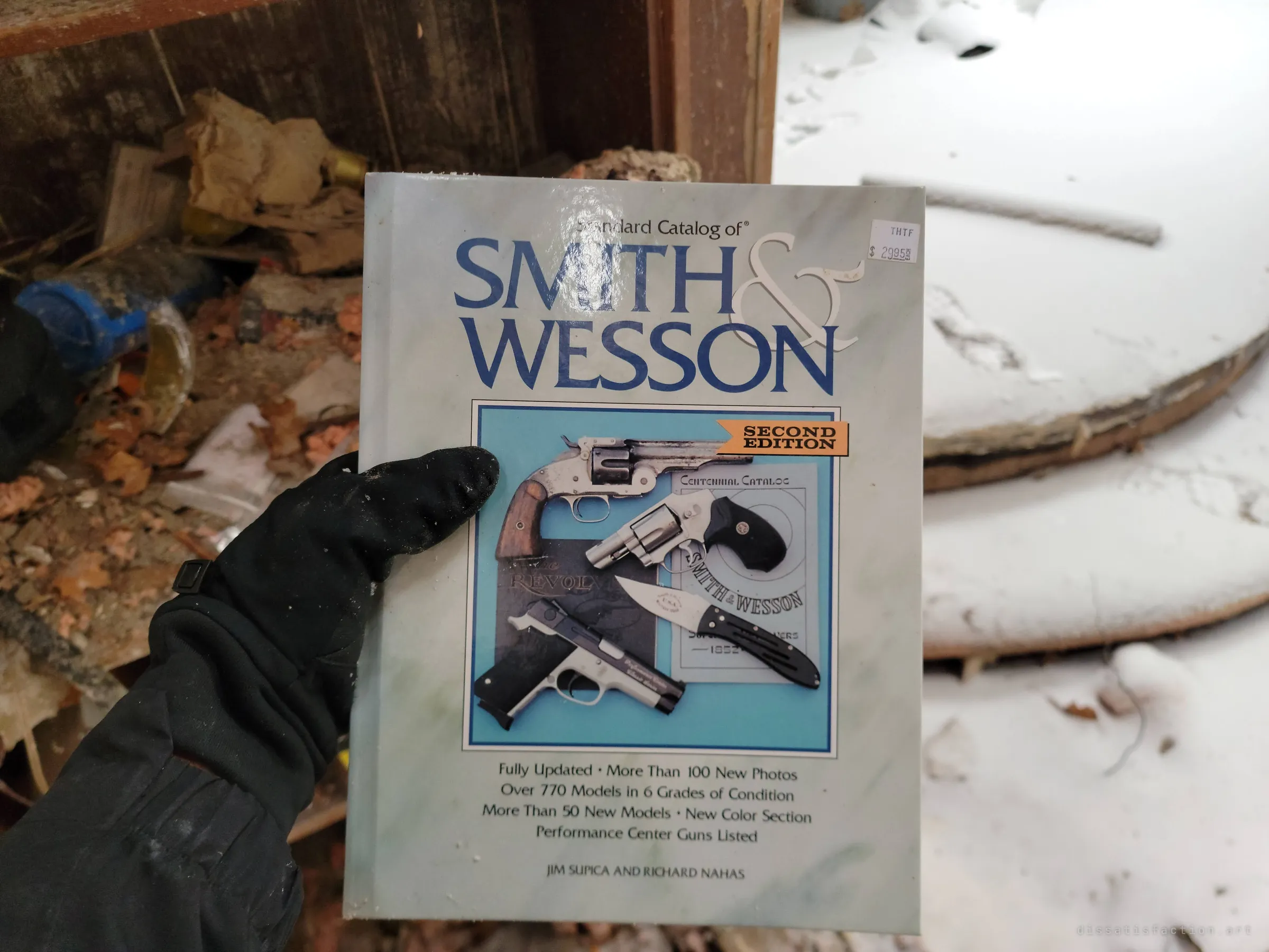 A gloved hand holds a collector's catalogue of revolvers and pistols above debris and snow-covered boards. Another find from the same shelf, fitting the album's prepper theme.