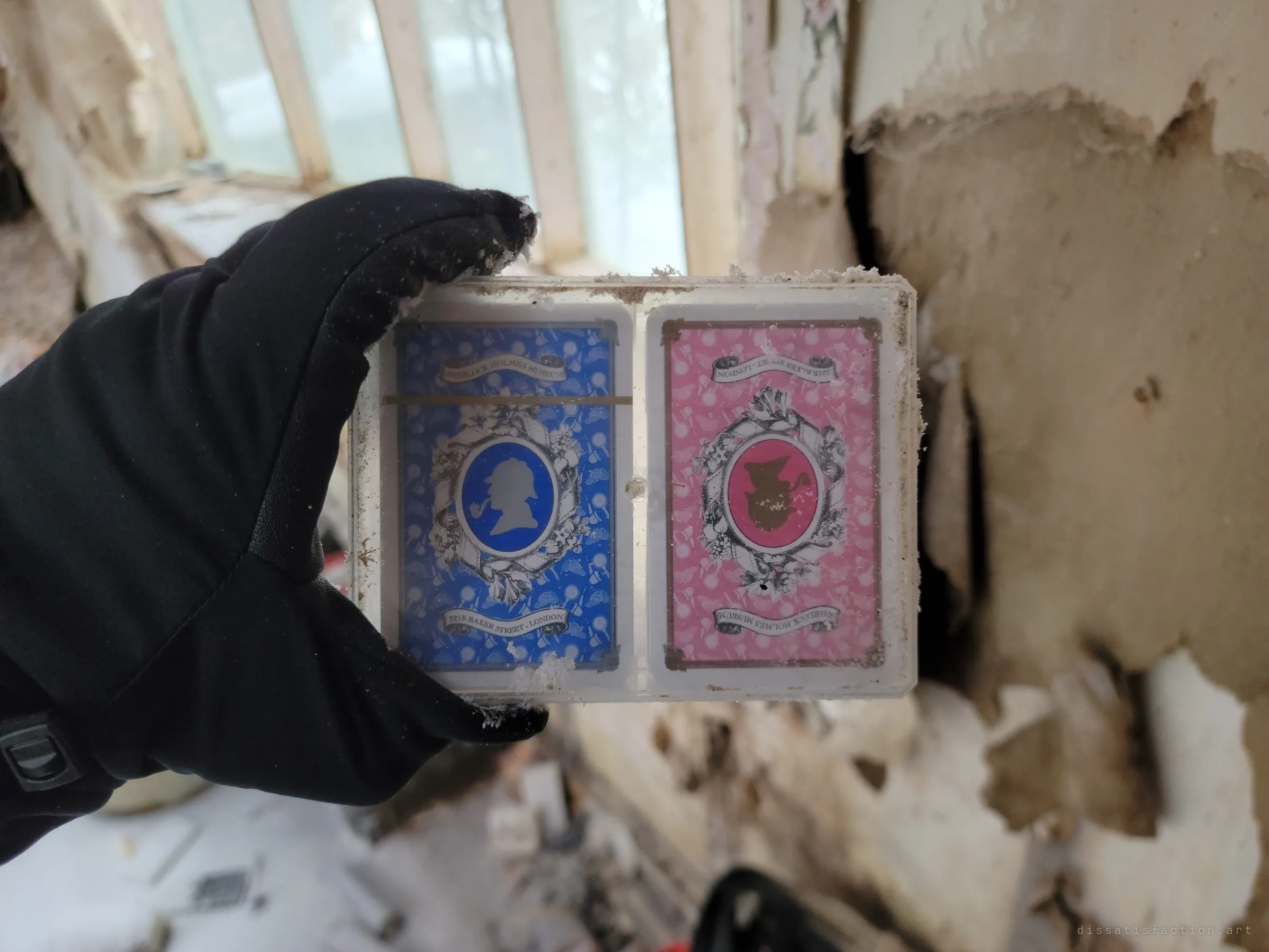 A gloved hand holds a dusty plastic case of two souvenir playing card decks, blue and pink, against peeling walls and a bright window. A tourist keepsake left behind in the wreckage.
