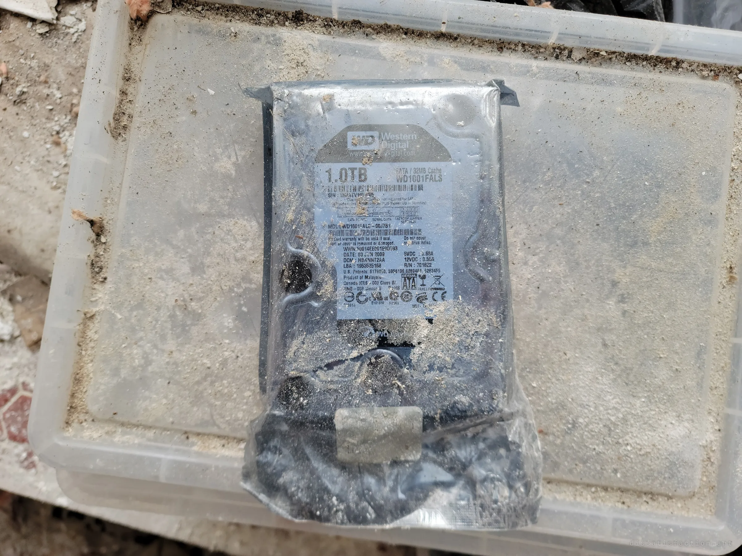 A sealed hard drive in its anti-static bag lies on a dusty plastic storage lid. Someone's data, still wrapped, left to the elements.