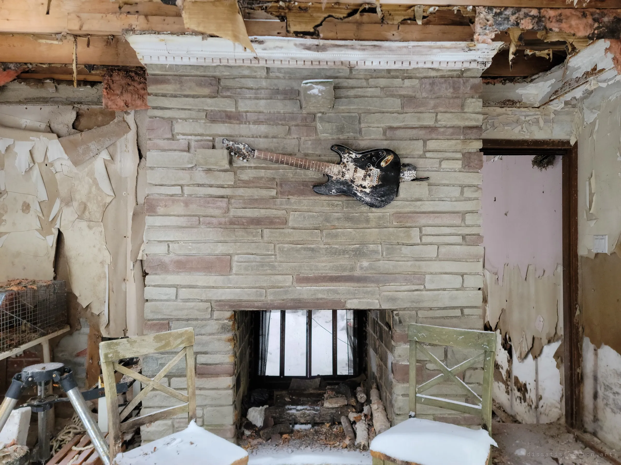 A wrecked electric guitar is mounted on a stacked-stone fireplace, flanked by two snow-dusted chairs under a torn-open ceiling. The through-fireplace frames snow outside, the centrepiece of the ruined living room.