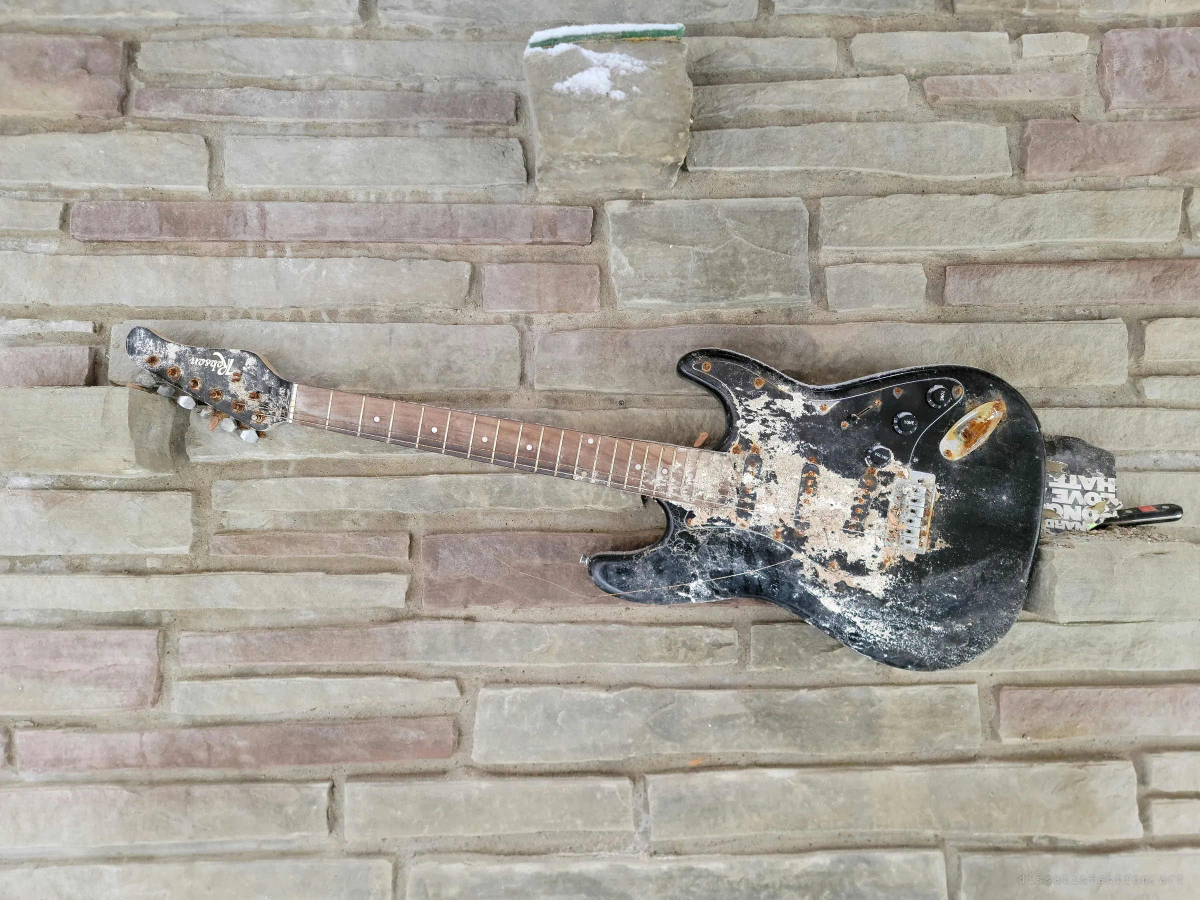 Close-up of the black electric guitar on the stone chimney, its body flaking and hardware rusted. A sticker tucked behind it adds a last bit of personality.