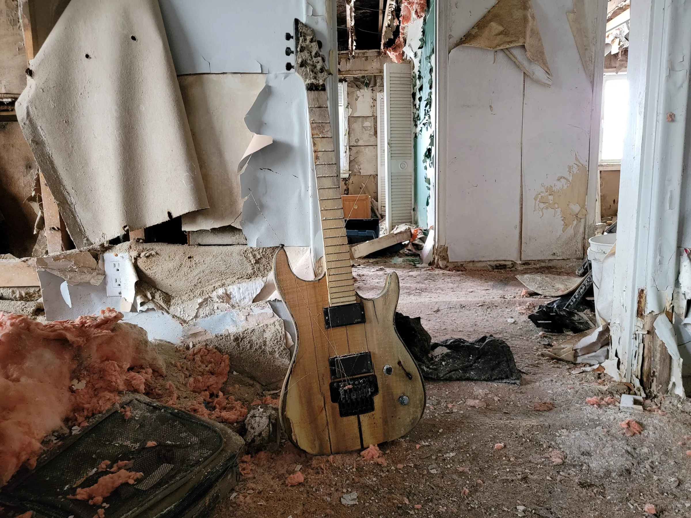 A cracked natural-wood electric guitar leans against a gutted wall amid pink insulation and plaster debris, with stripped rooms beyond. Another instrument abandoned in the house.