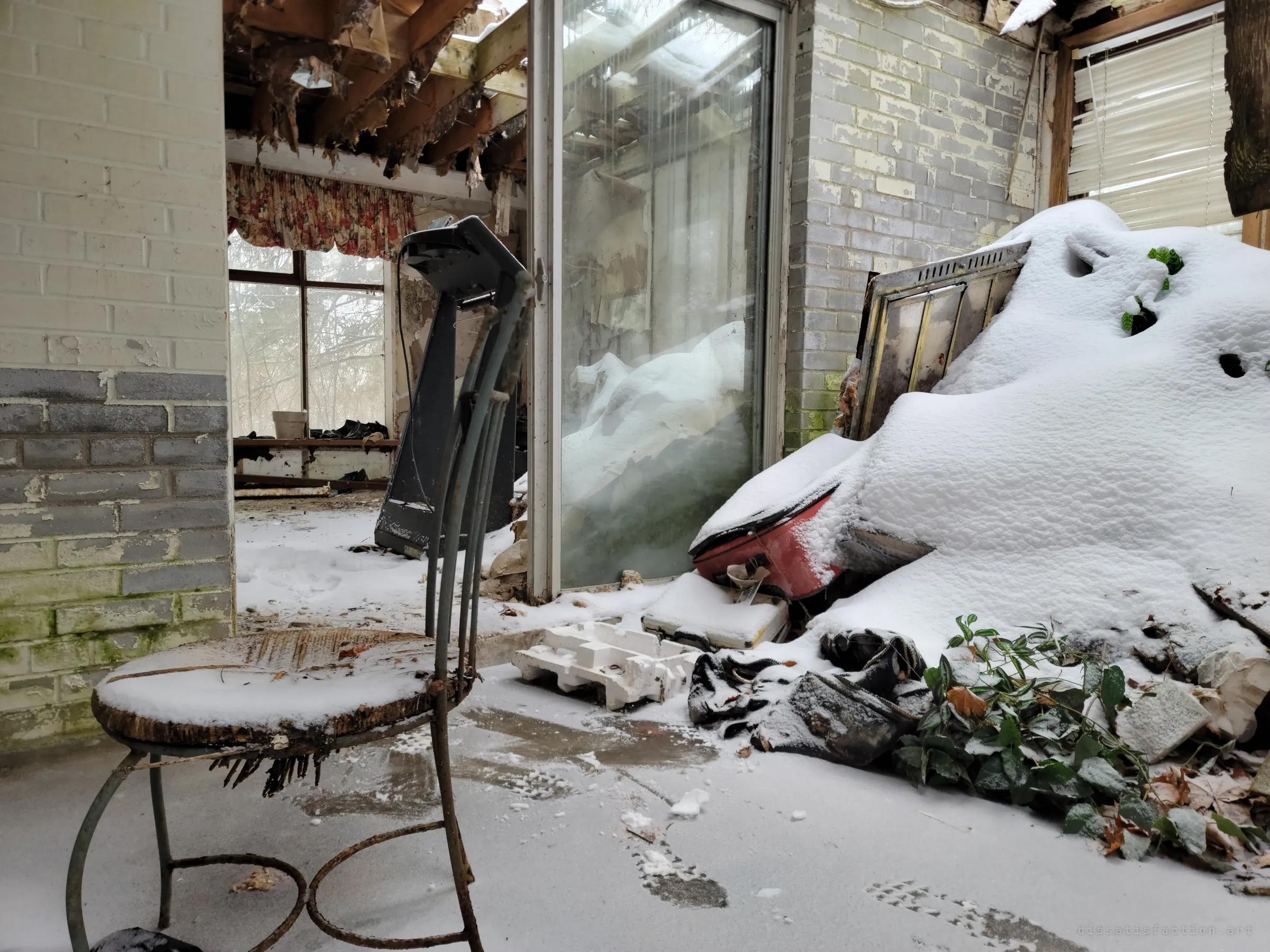A rusted bistro chair sits on a snow-covered floor beside a collapsed ceiling, a treadmill and heaped, snow-buried junk. Winter has moved inside the house.