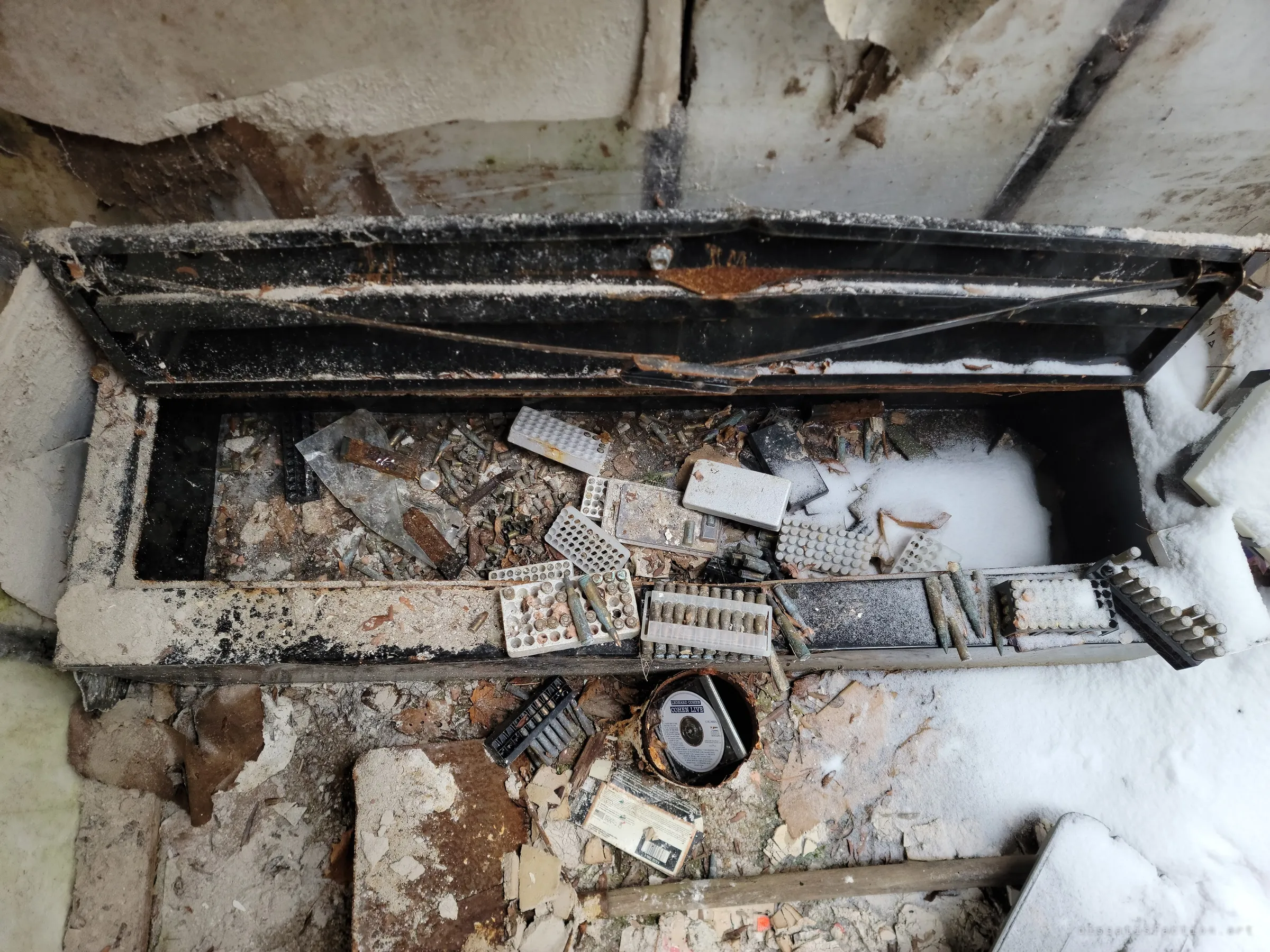 A long black metal chest dusted with snow, full of loose brass casings, cartridges and plastic ammo trays, with a rusted tin and a music CD on the floor below. It shows how much ammunition was stockpiled and left to the weather.