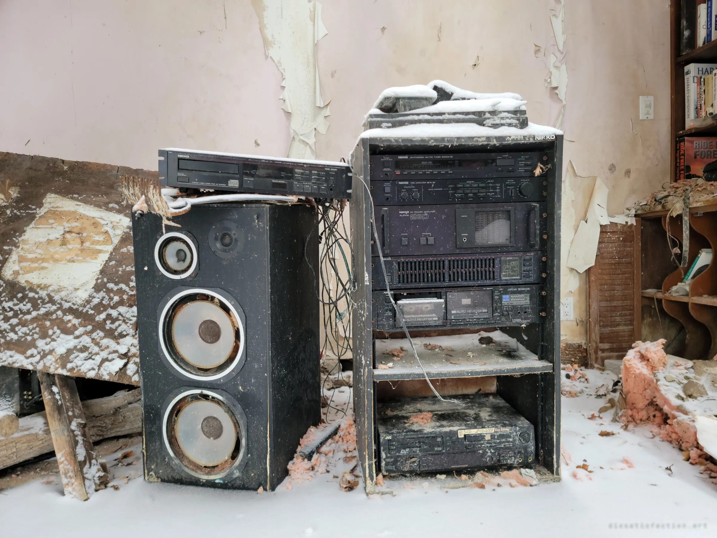 A tall floor speaker and a rack of 1980s hi-fi separates stand in snow and pink insulation, under peeling pink wallpaper beside a bookcase. A once-cared-for stereo setup slowly buried indoors.