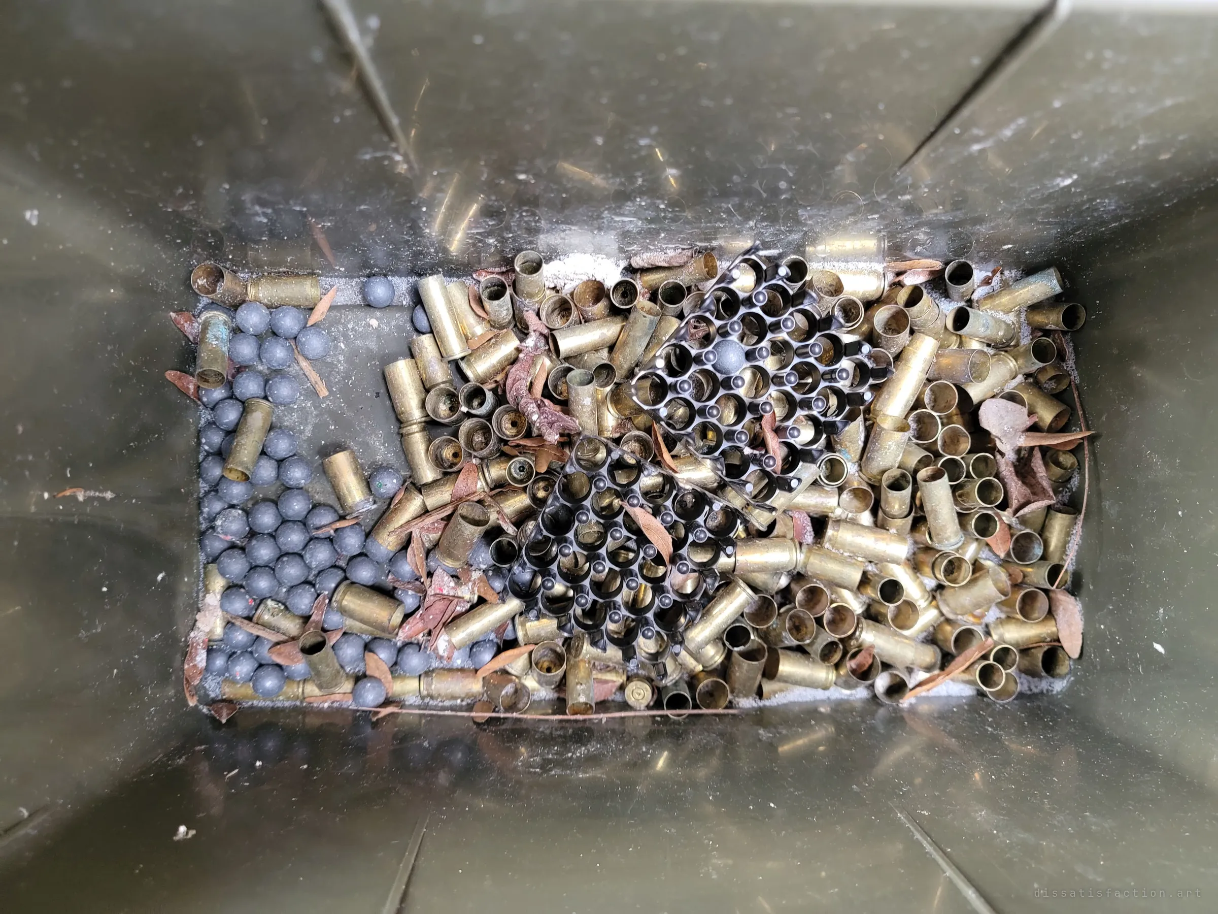 Looking down into an olive ammo can holding spent brass casings, lead balls and black primer trays mixed with dry leaves. Reloading scraps kept long after the shooting stopped.